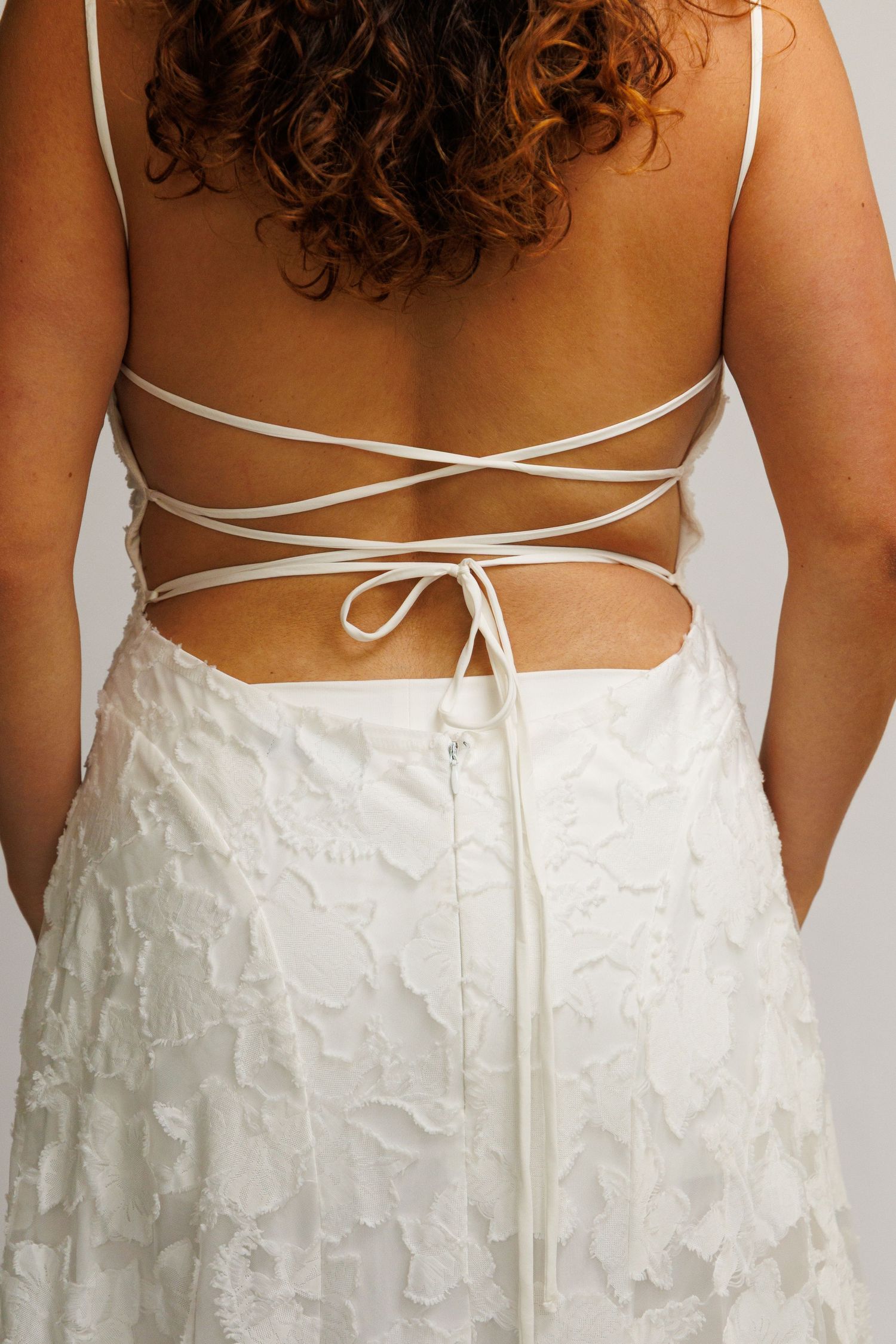 Woman wearing a white backless dress with criss-cross tie straps and textured floral skirt, viewed from behind.