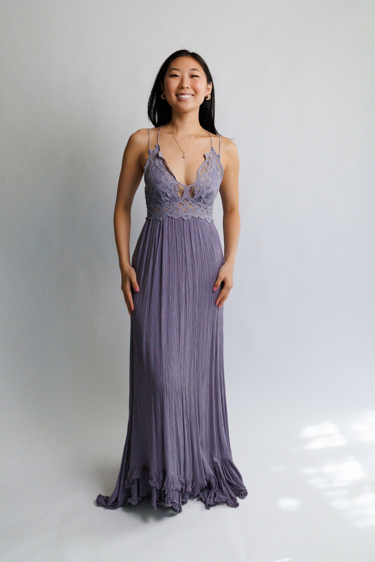 Woman in lavender lace bodice maxi dress with spaghetti straps and pleated skirt, smiling in studio.