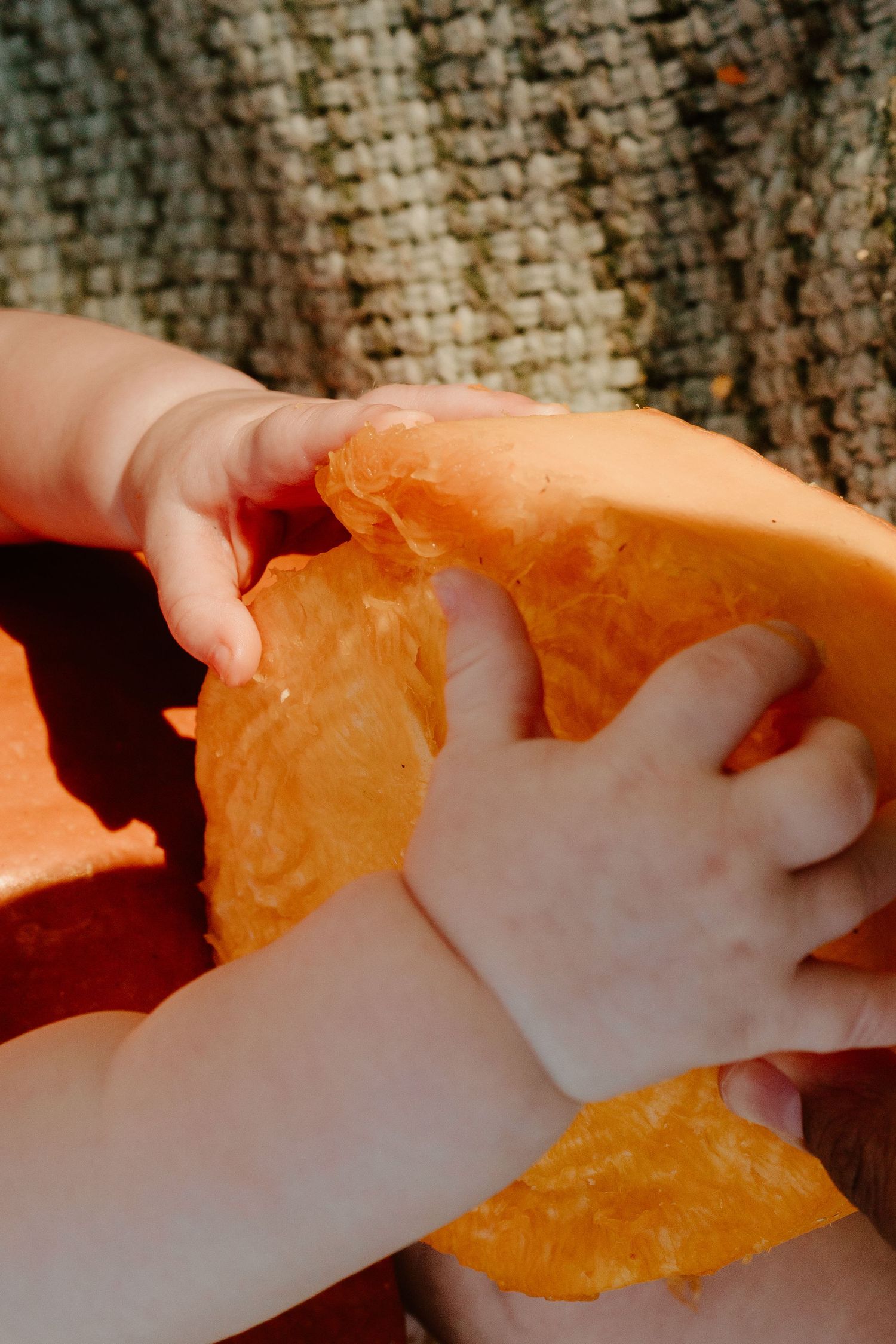 Small child's hands gripping and exploring a hollowed-out orange pumpkin half outdoors, set against a woven textured background.