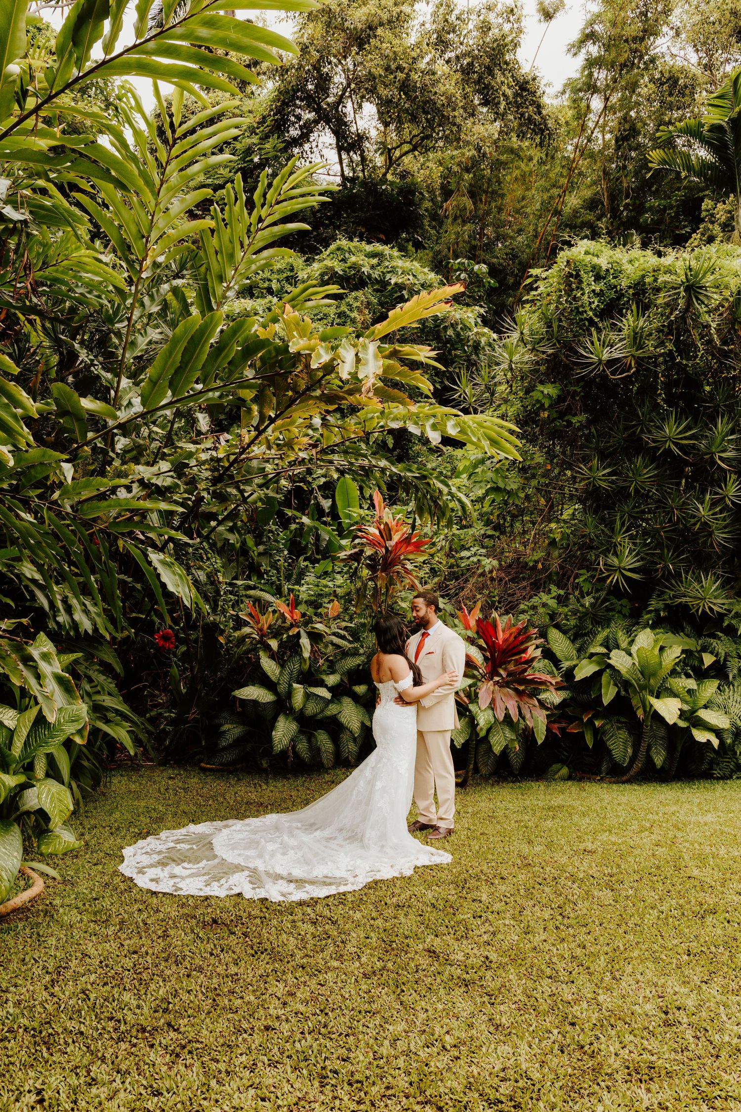 Bride and groom embrace in a lush tropical garden, surrounded by vibrant green foliage and red plants.
