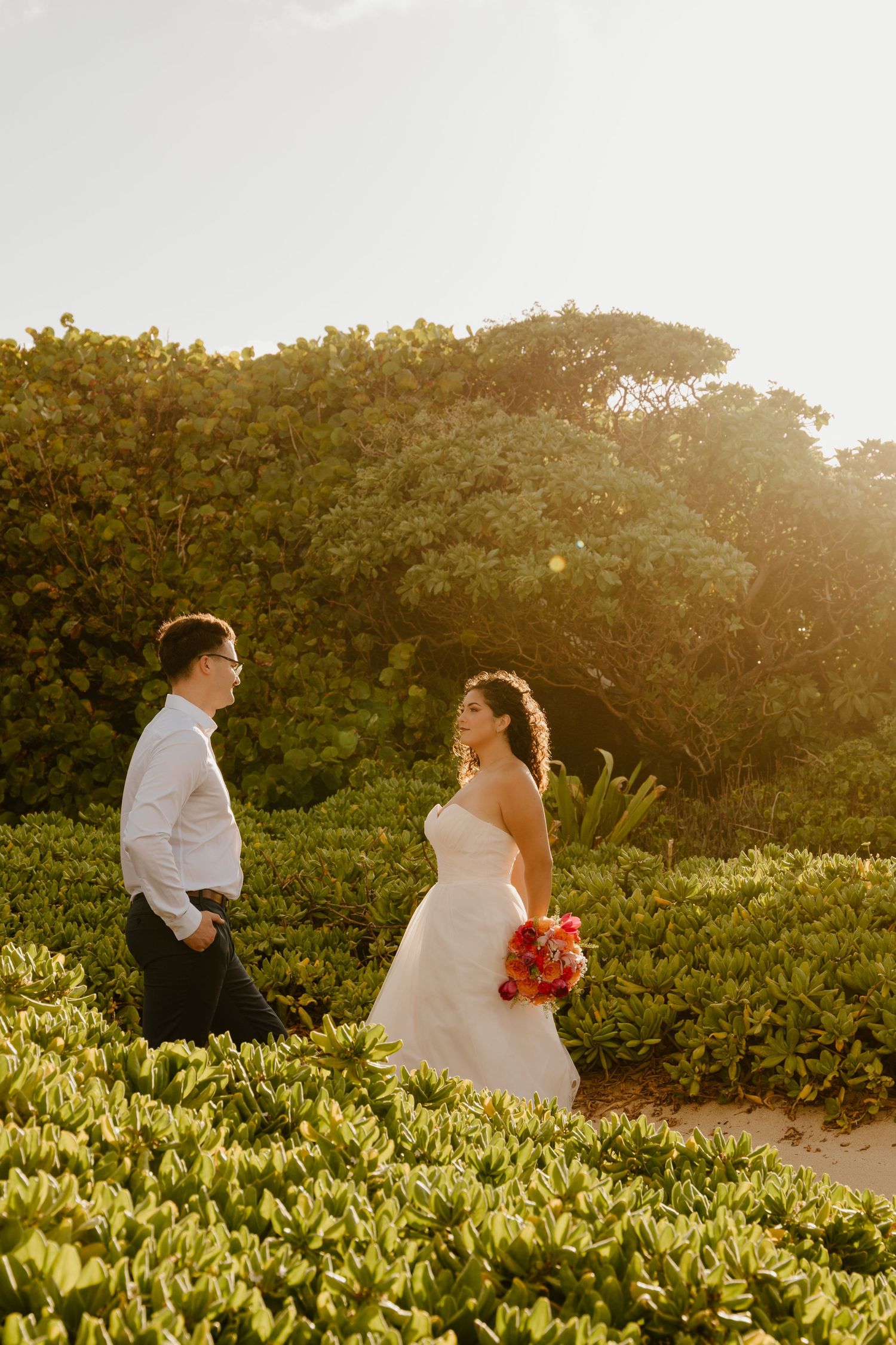 Bride and groom share a romantic moment surrounded by lush greenery and golden sunlight during outdoor beach wedding.