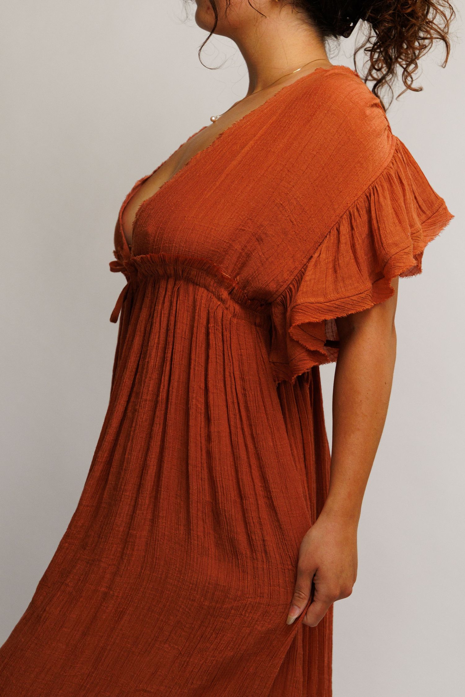Woman wearing a rust orange flutter sleeve empire waist maxi dress with deep V-neckline and textured fabric.