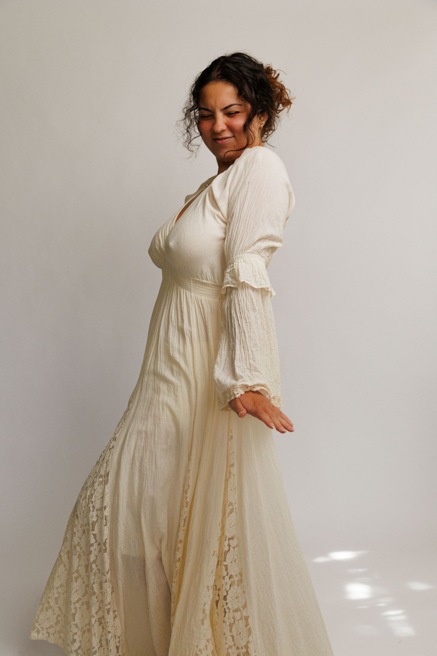 Woman in ivory lace maxi dress with long sleeves, smiling over shoulder, elegant bridal style gown on white background.