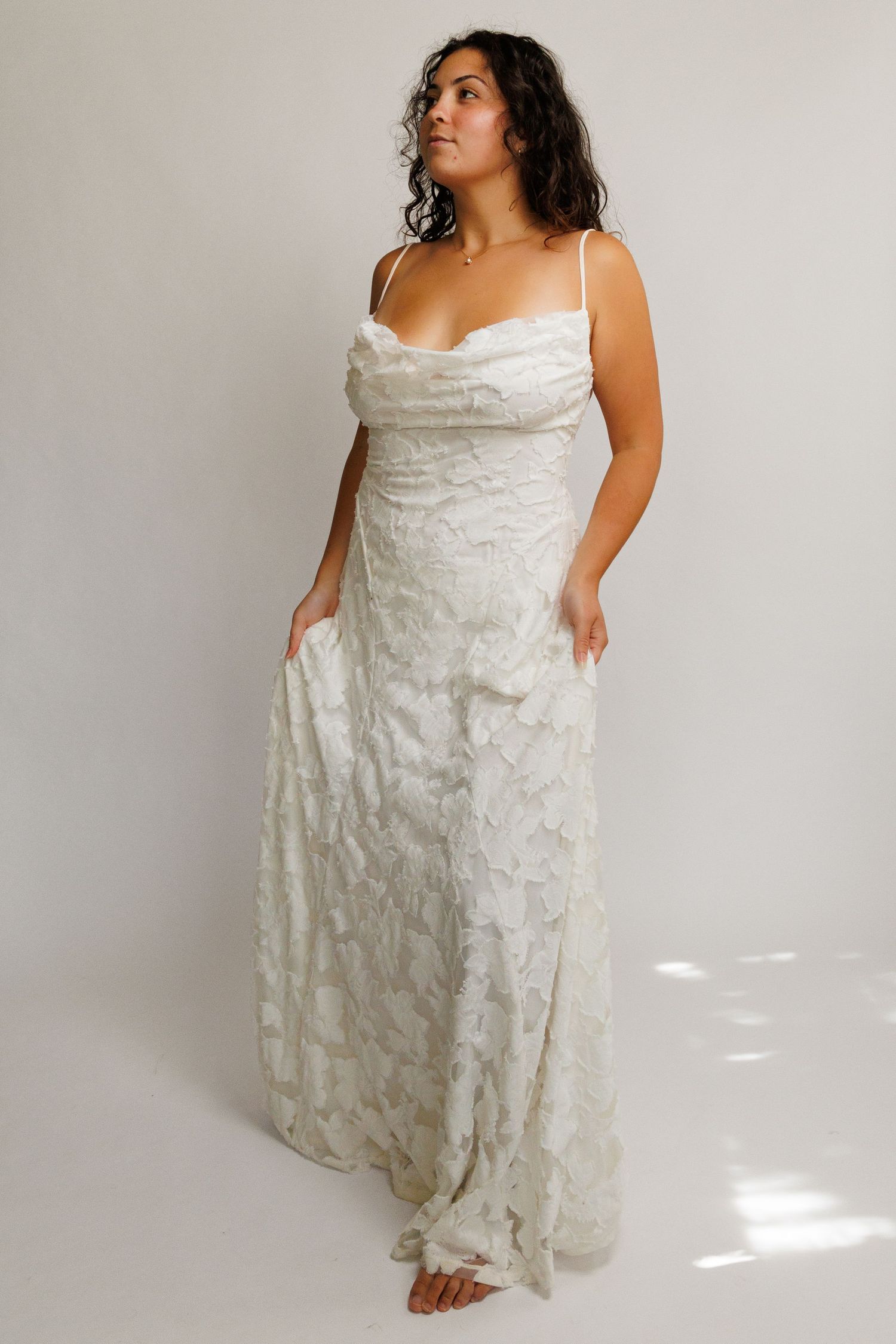 Woman wearing a white floral textured spaghetti strap maxi dress with cowl neckline on light background.