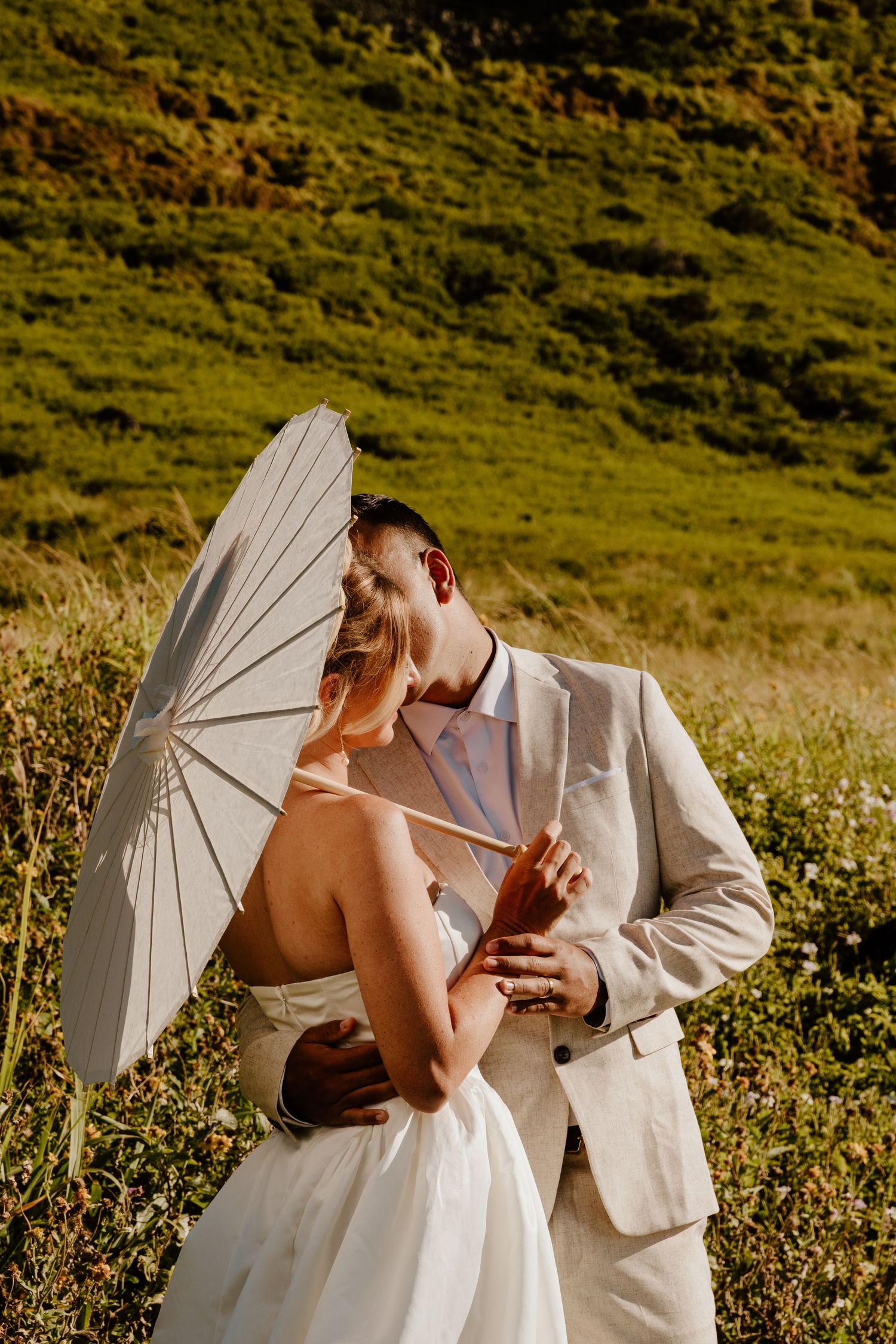 Bride and groom sharing romantic moment under white parasol in sun-drenched green hillside field.