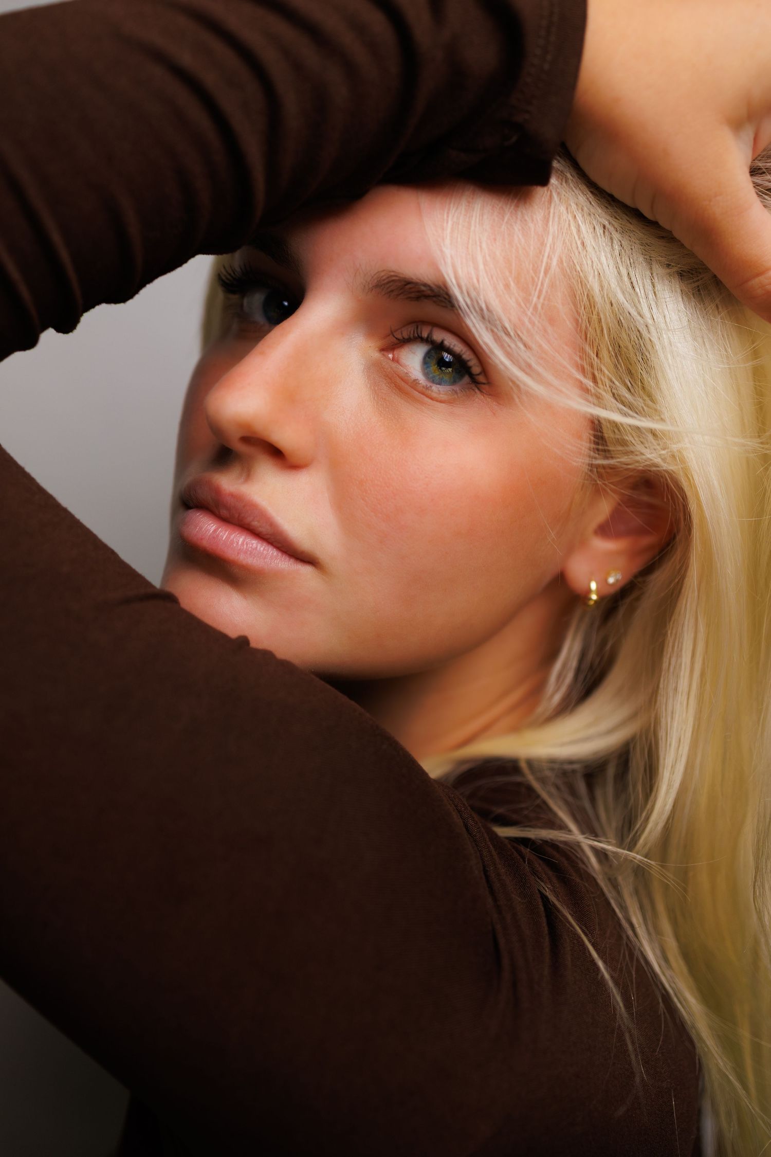 Young blonde woman in brown top with hands raised near her head, gazing upward in a close-up portrait shot.