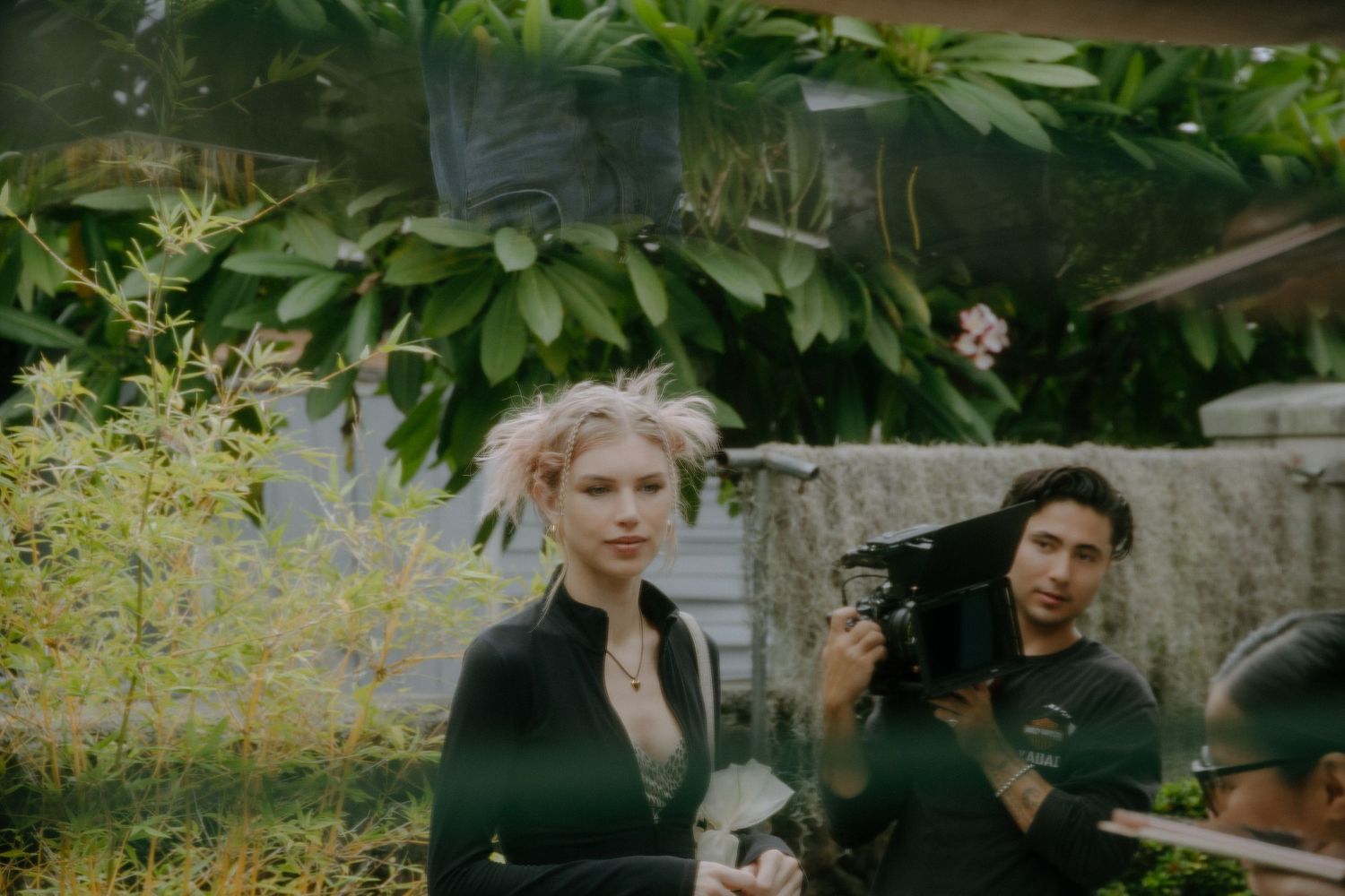 Blonde woman in black jacket stands near lush green plants while man holds vintage camera outdoors.