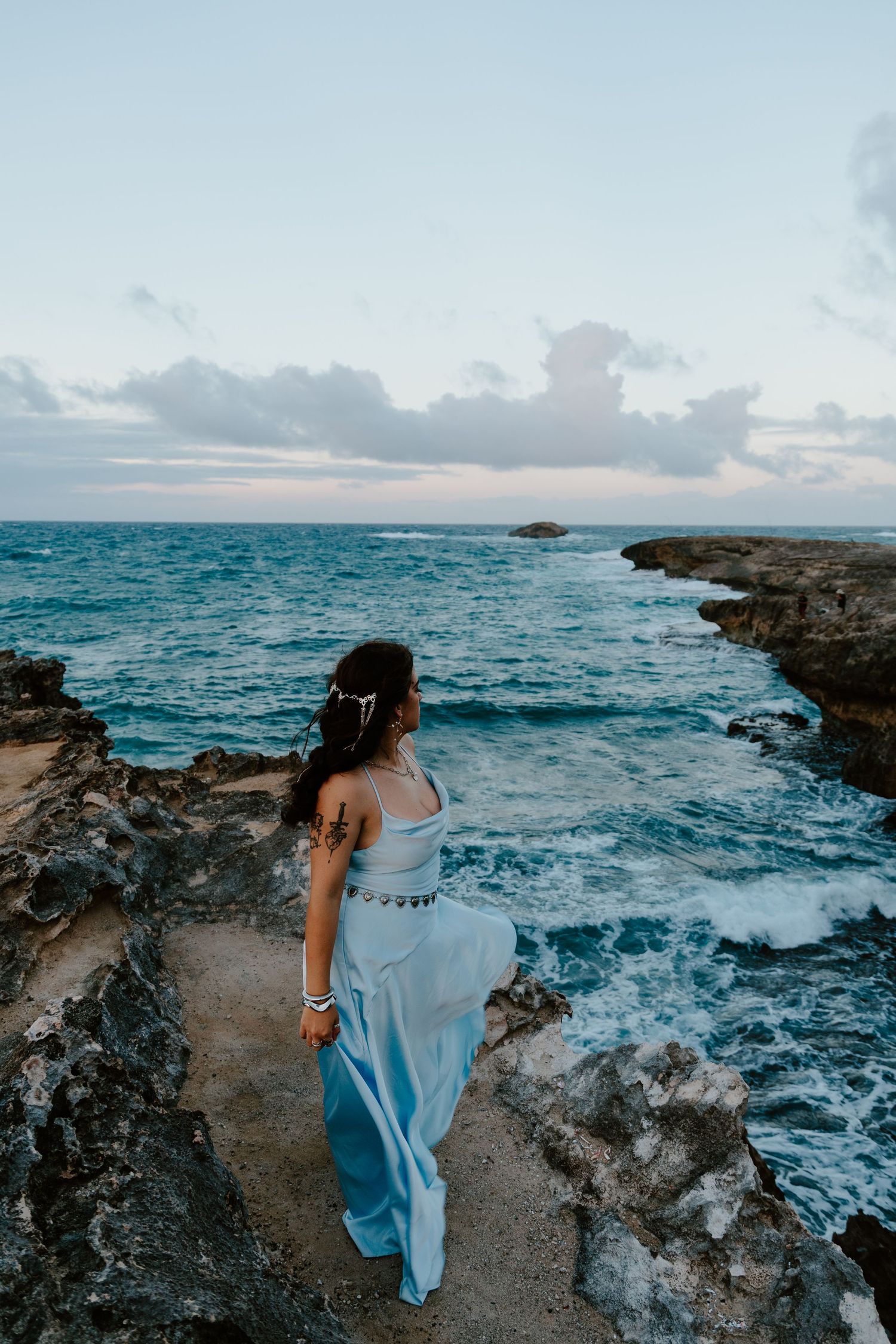 Woman in flowing blue dress stands on rocky ocean cliffs at sunset, gazing at turquoise waves below.