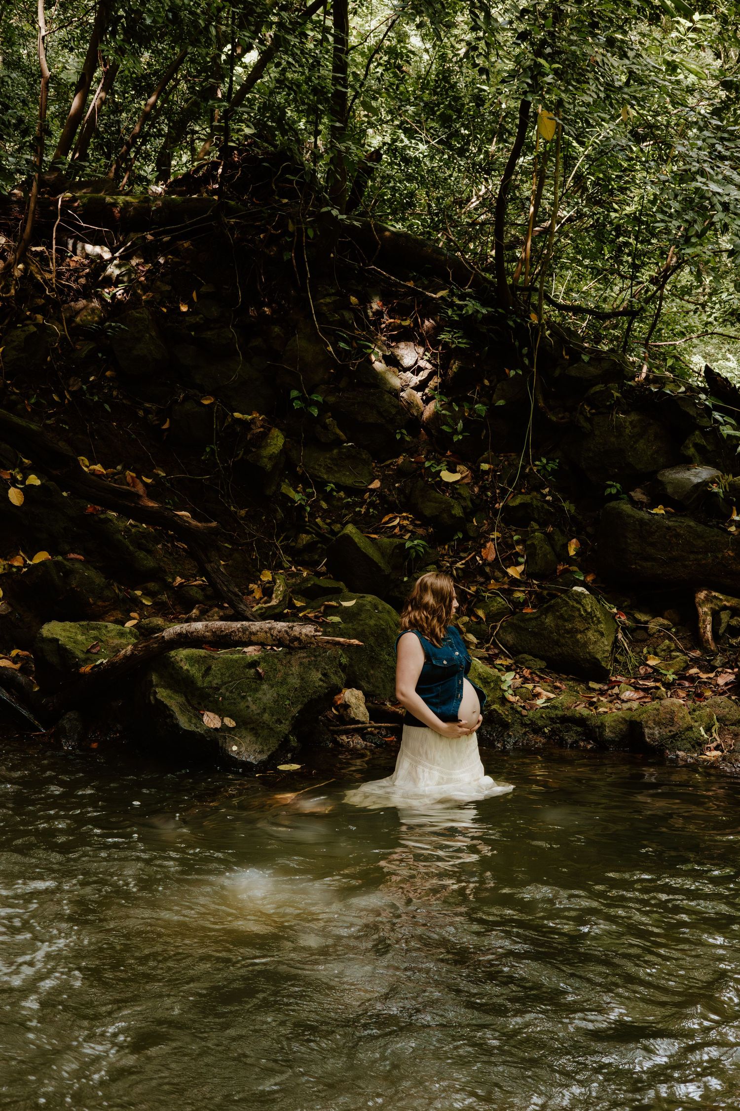 Pregnant woman in denim vest wading through forest creek surrounded by lush green trees.