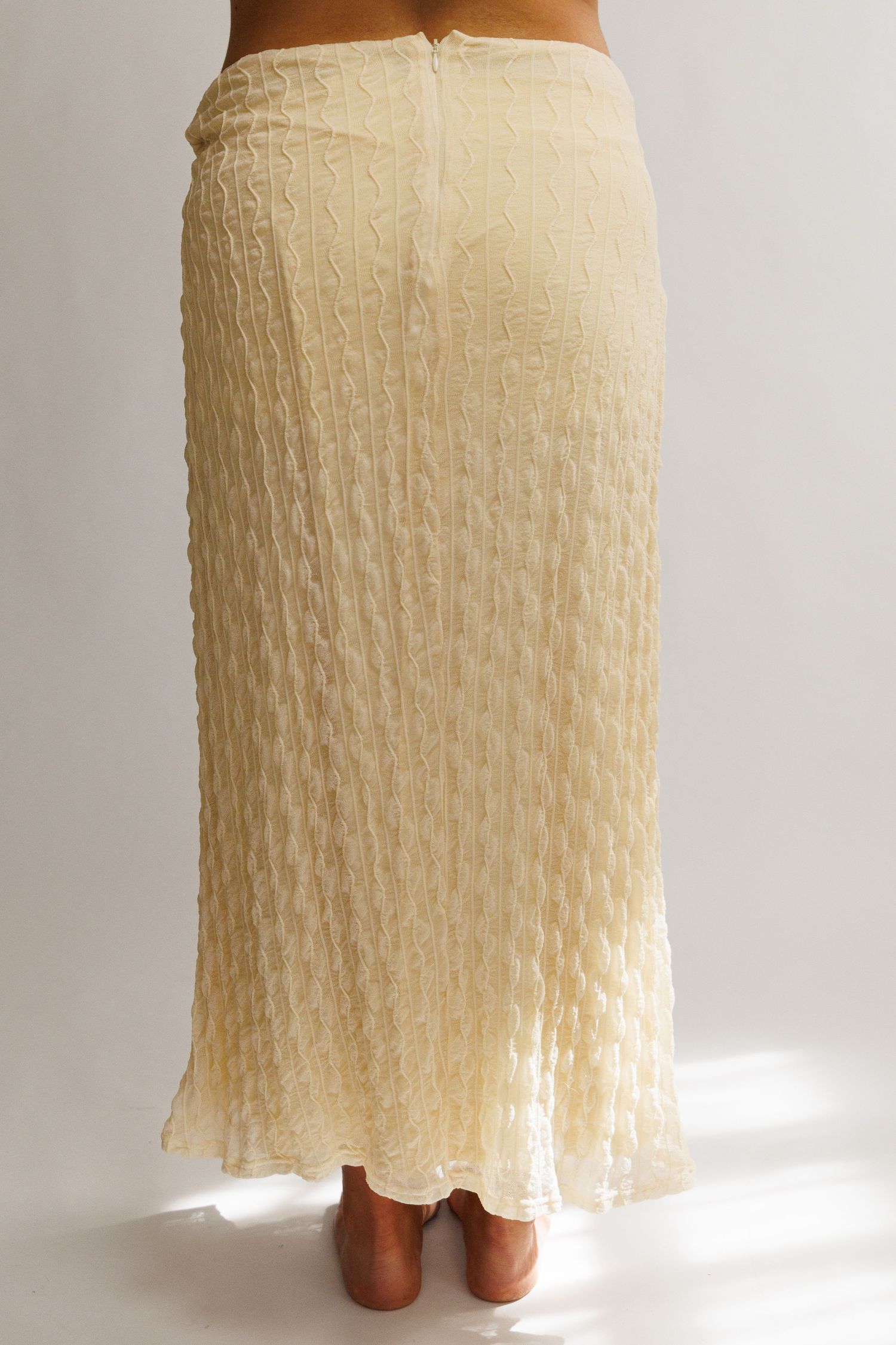 Back view of a cream textured maxi skirt with crinkled fabric and a fitted waistband, shown on a model.