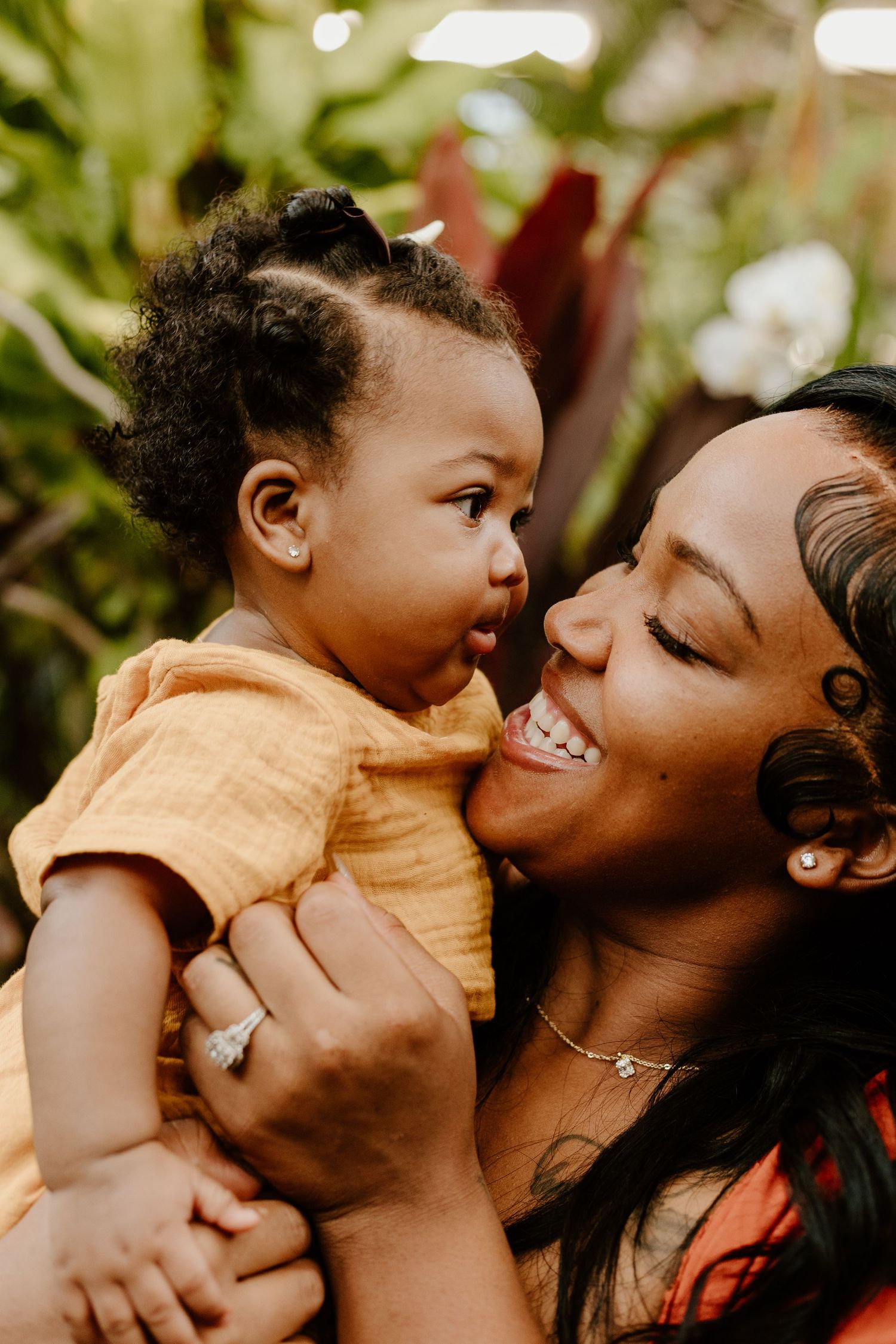 A joyful mother laughs while holding her adorable baby in a yellow outfit outdoors.
