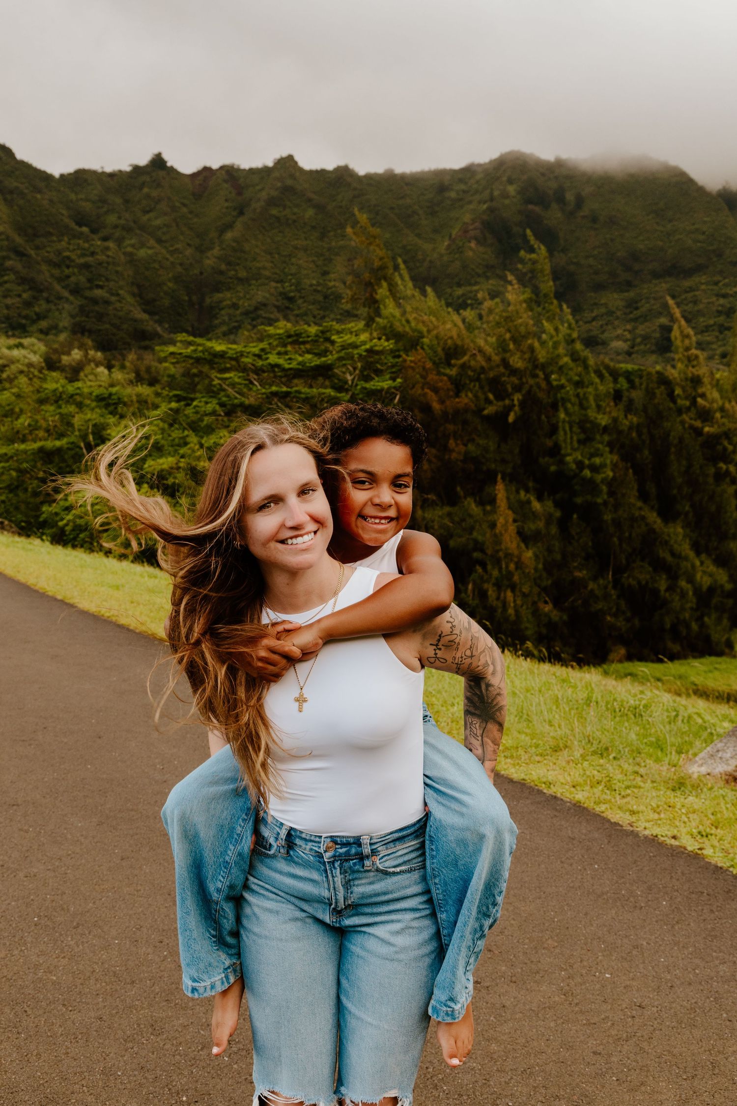 Two women smiling, one giving the other a piggyback ride on a road with green mountains.