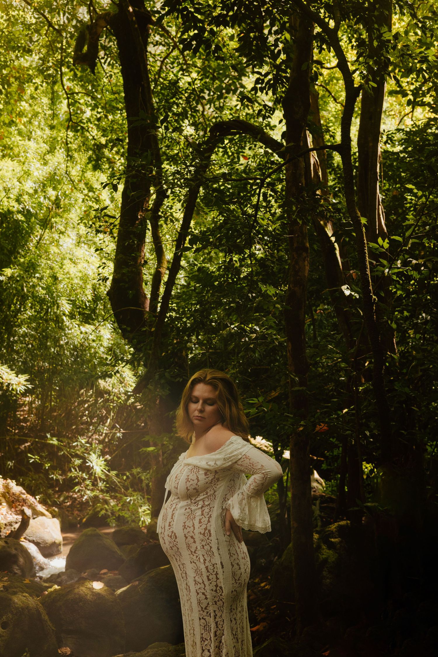 Pregnant woman in white lace off-shoulder gown standing in sunlit forest with green trees.
