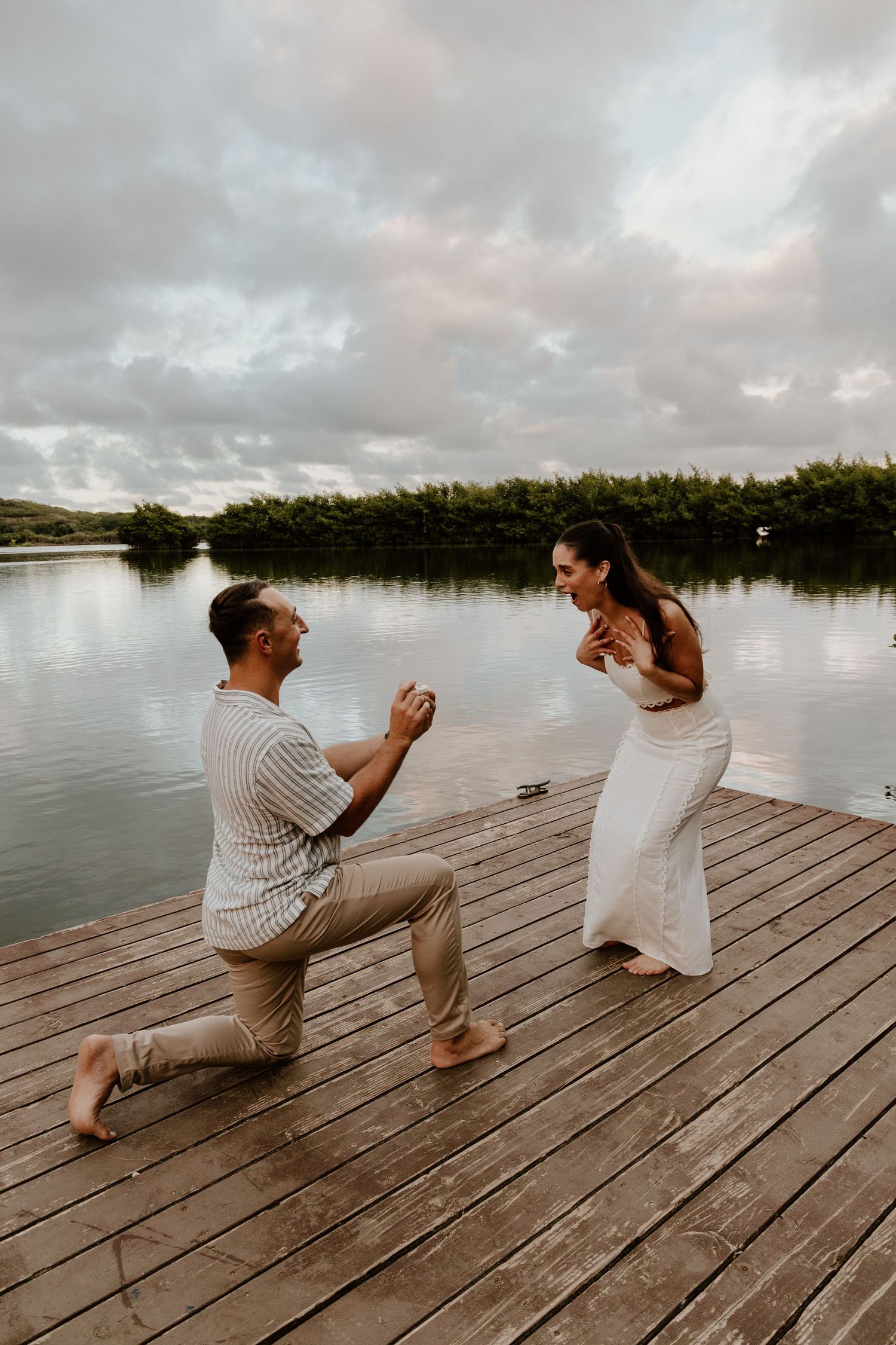 Man proposing on one knee to surprised woman on wooden dock at sunset over calm lake.