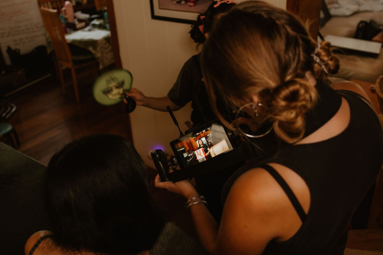 Two women looking at a camera screen together indoors, one holding a cucumber slice during a behind-the-scenes moment.