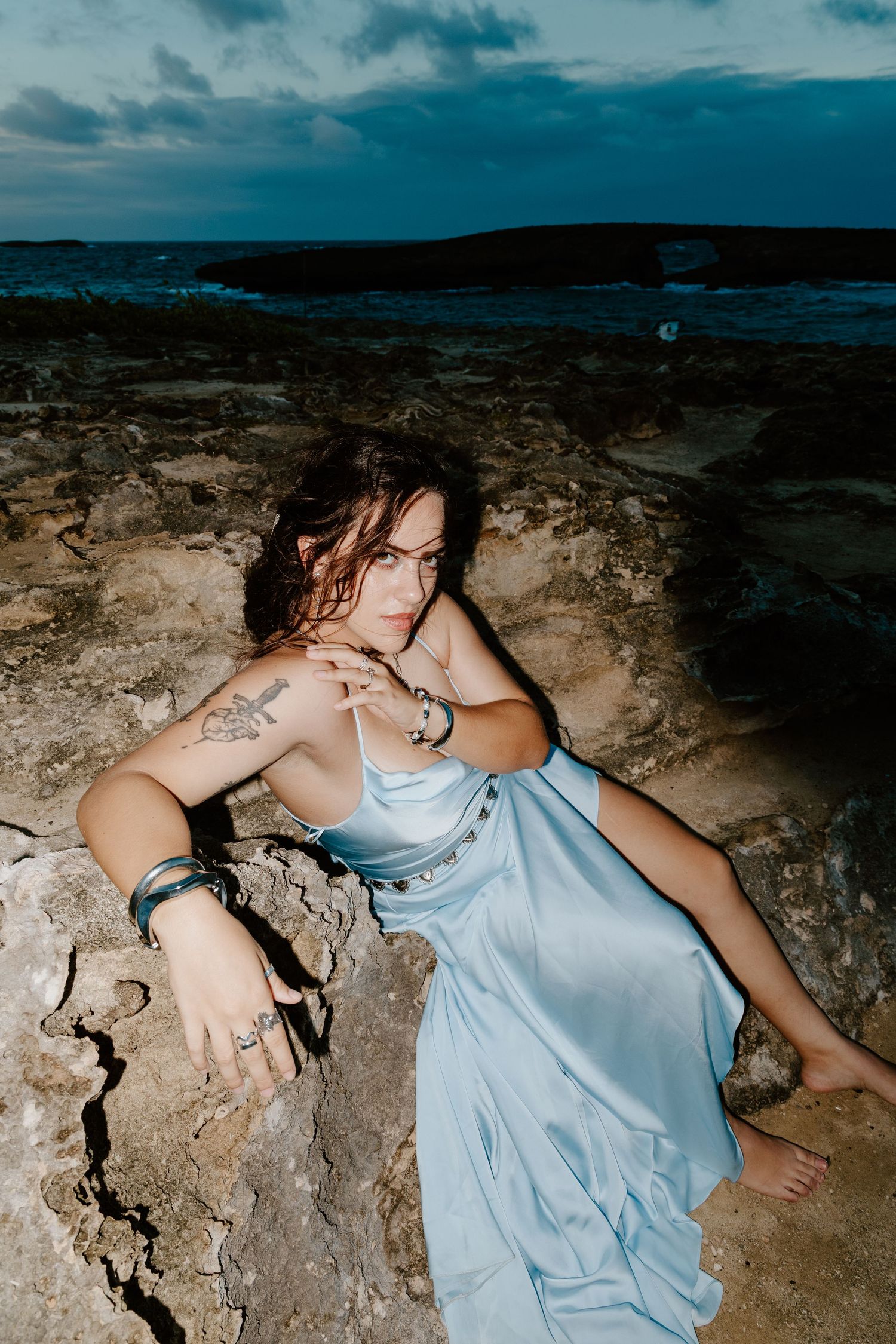 Woman in blue satin dress posing on rocks at dusk with ocean backdrop.