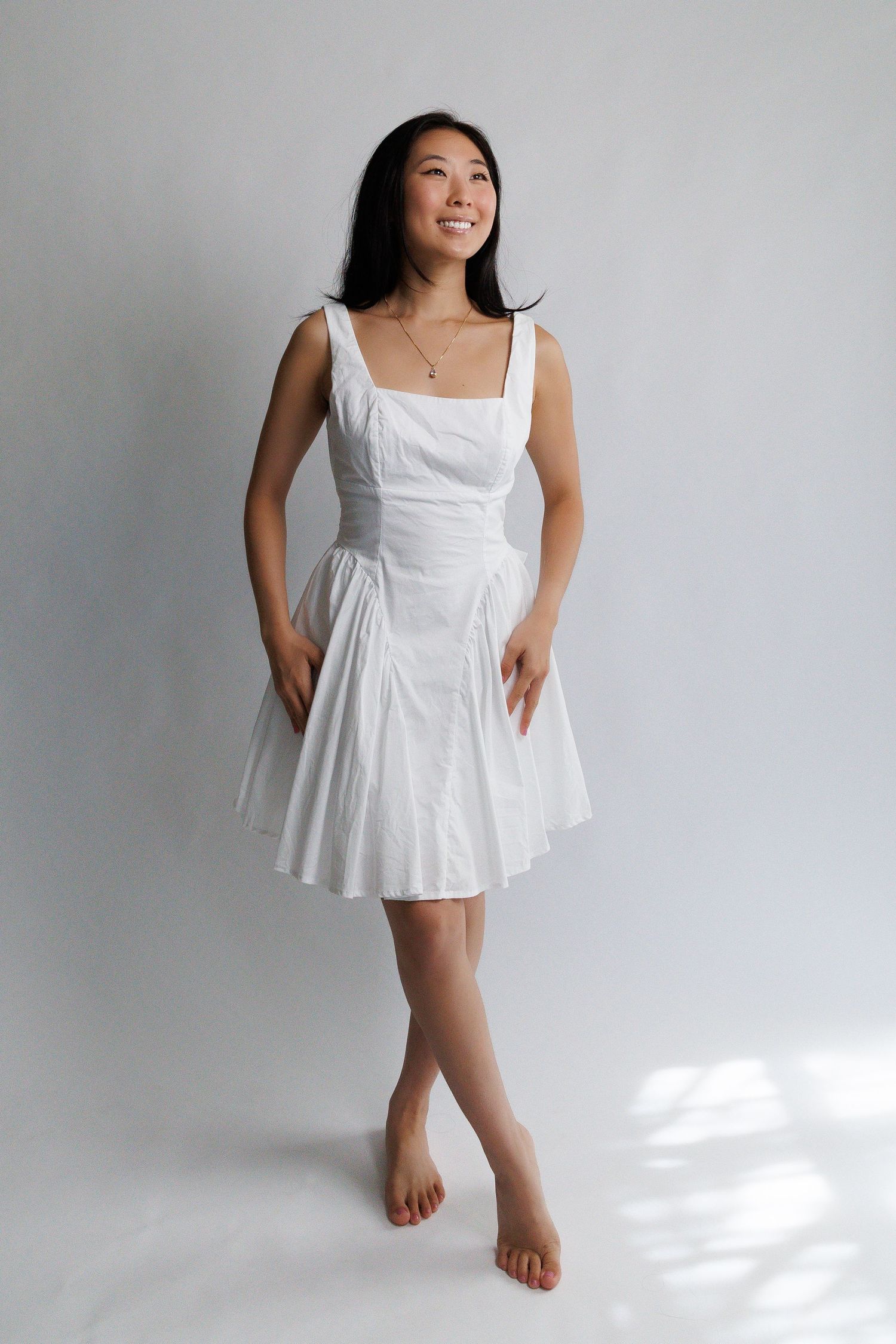Smiling woman wearing a white sleeveless fit-and-flare mini dress, posing barefoot against a light gray background.