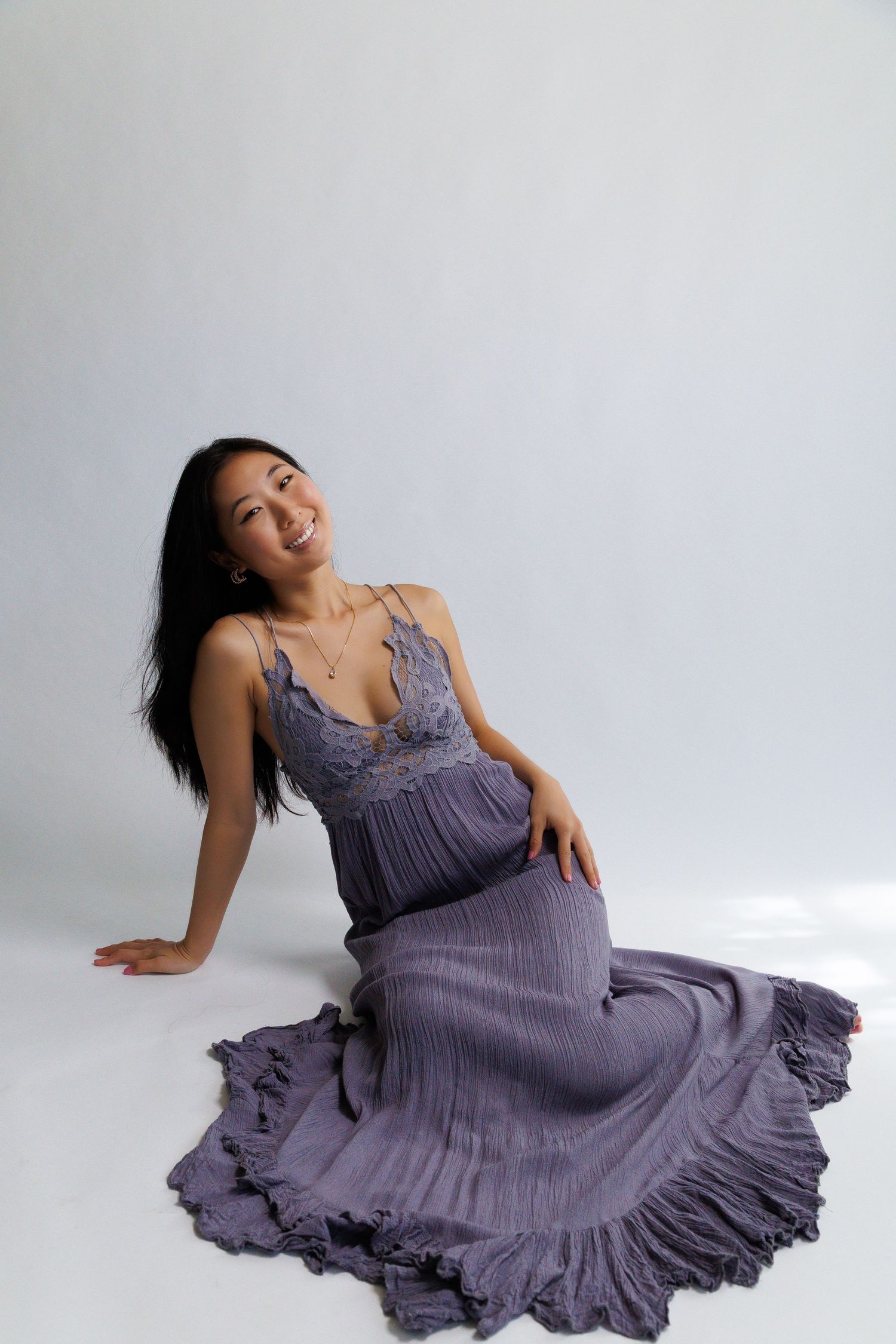 Smiling woman in purple maxi dress sitting gracefully on white studio floor with long dark hair.
