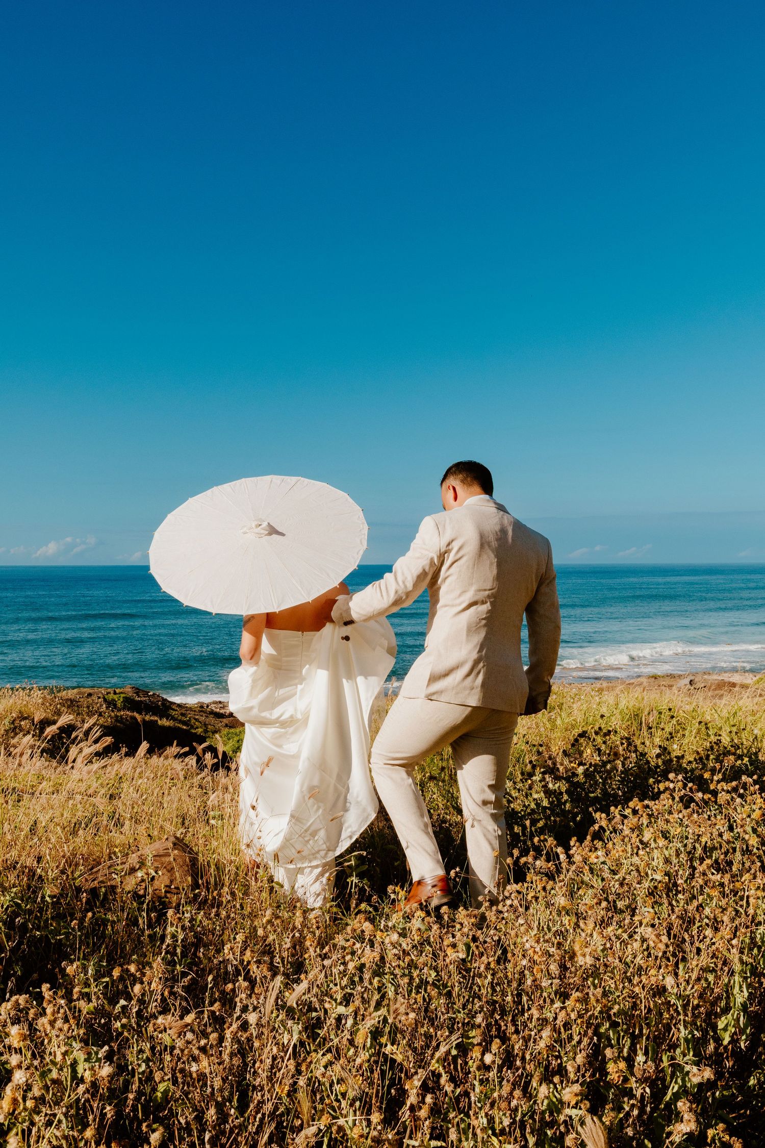 Bride and groom walking toward ocean with white parasol umbrella on sunny coastal cliffside.