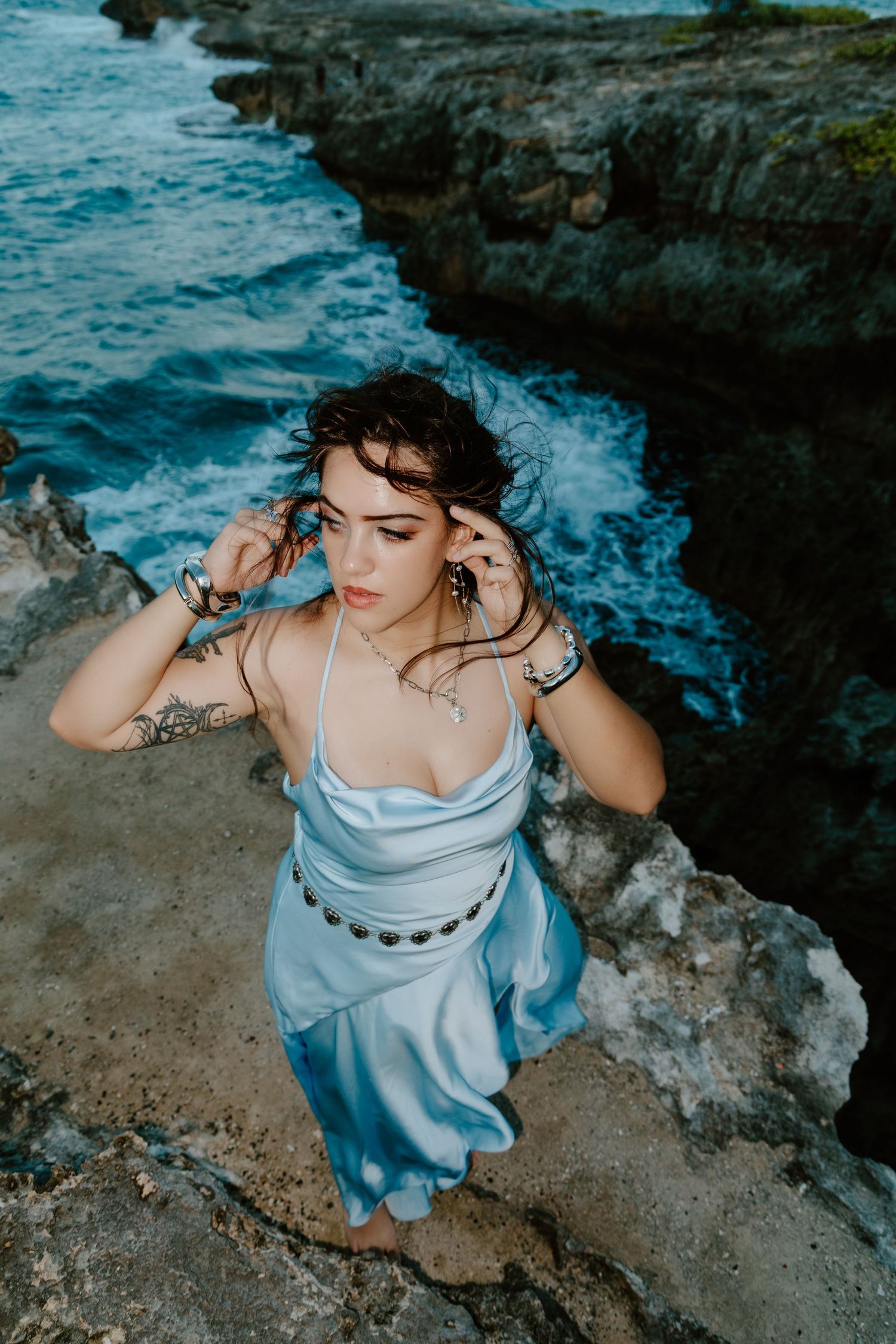 Woman in blue satin dress sitting on rocky coastal cliffs with ocean waves, wearing silver jewelry and tattoos.