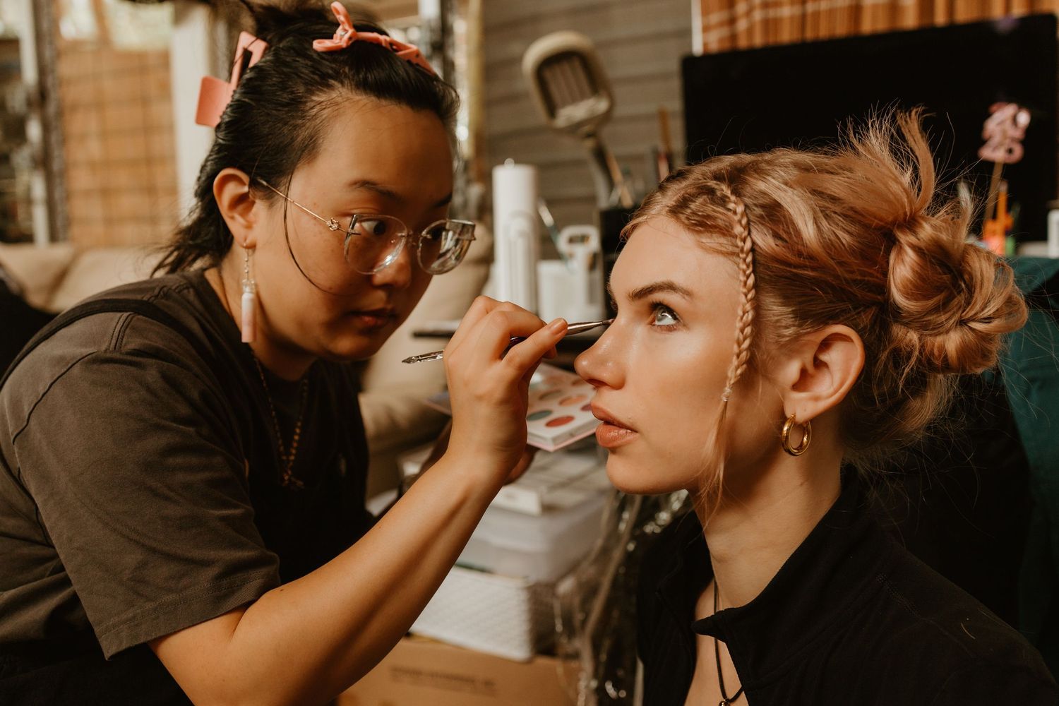 Makeup artist applying eye makeup to a young woman with an elegant updo hairstyle and gold accessories.