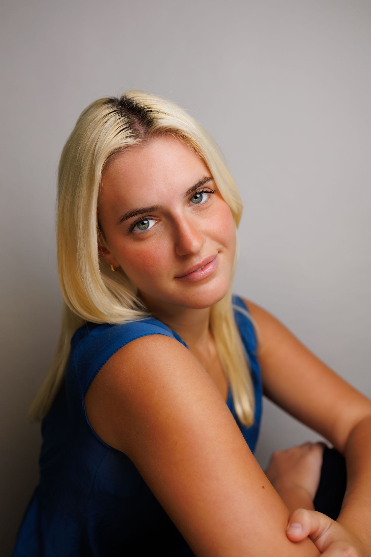 Smiling blonde woman in a blue sleeveless top poses confidently against a gray background.