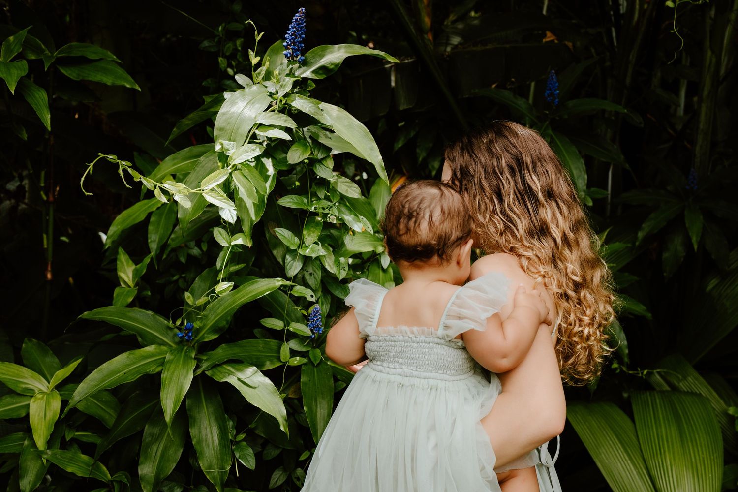 Two young girls in light blue dresses hugging among lush tropical green plants with blue flowers.
