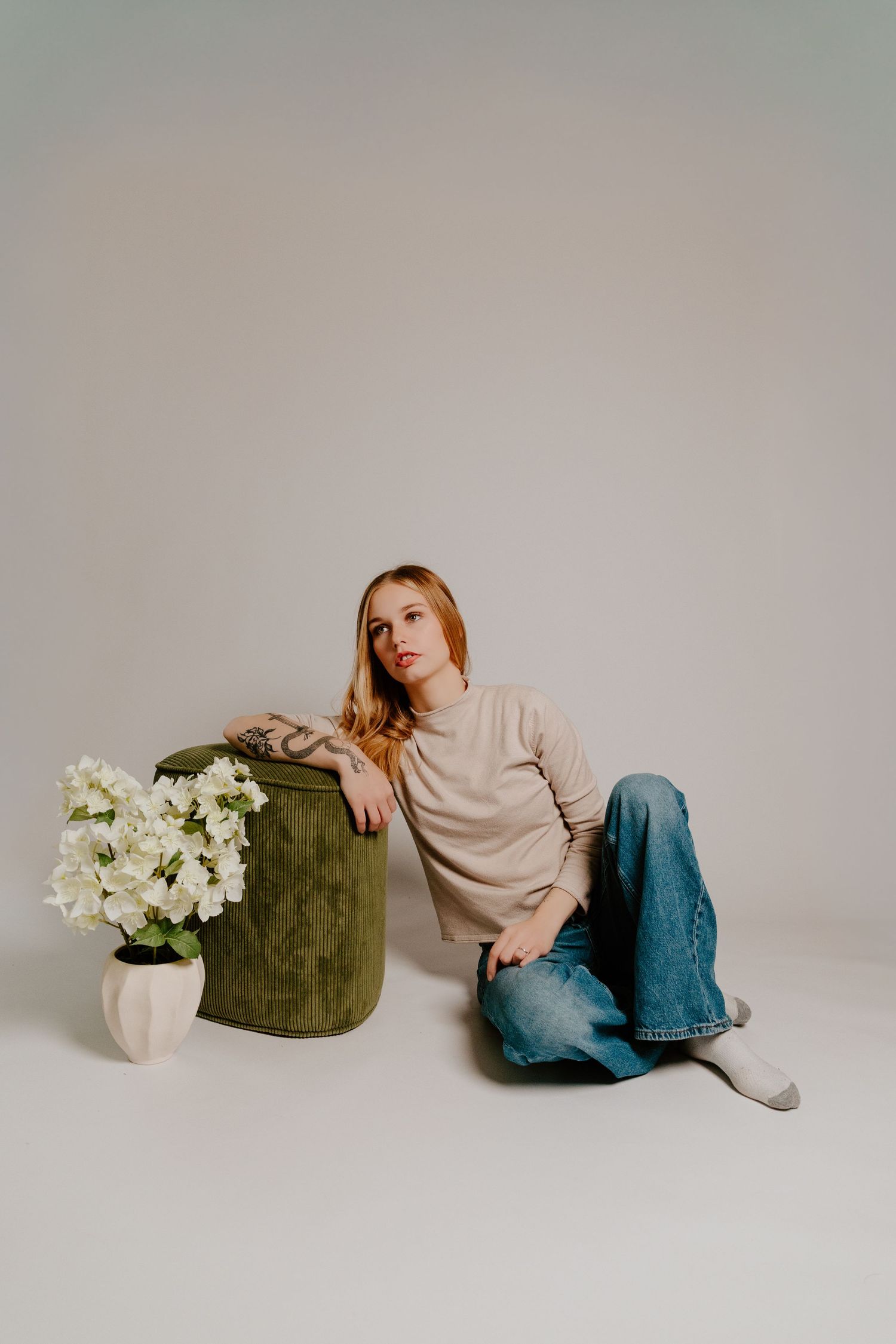 Blonde woman with tattoos sitting on floor beside green ottoman and white flowers in neutral studio setting.