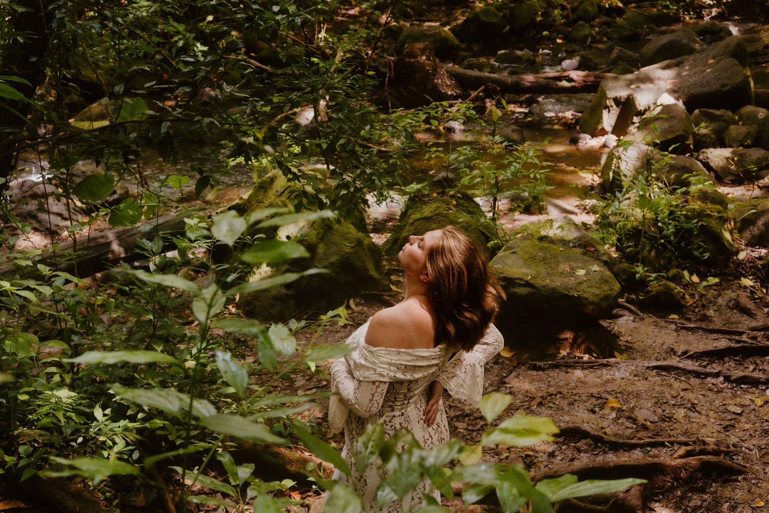 Pregnant woman in off-shoulder lace dress sitting among lush green foliage in forest setting.