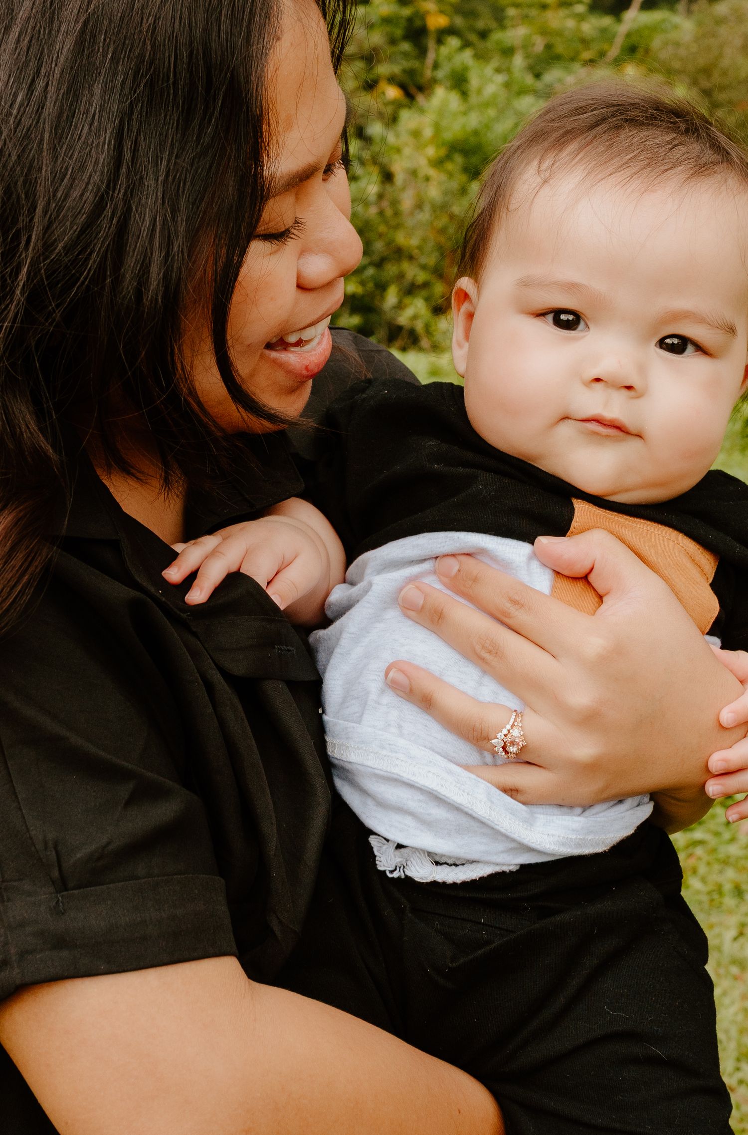 Mother in black outfit tenderly holding chubby baby outdoors with green foliage background.
