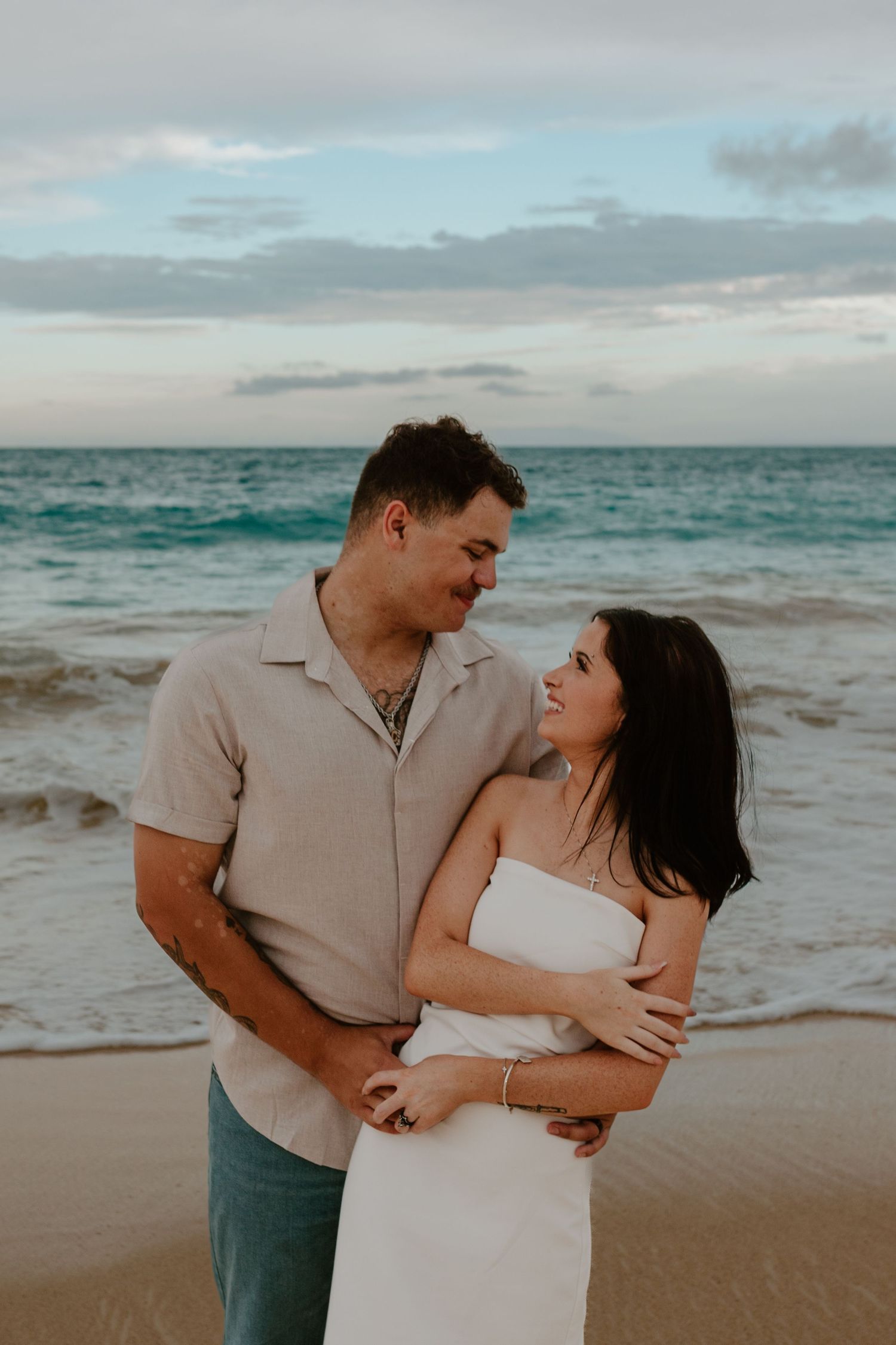 Couple gazing at each other lovingly on a beach at sunset, woman in white dress held by man in beige shirt.