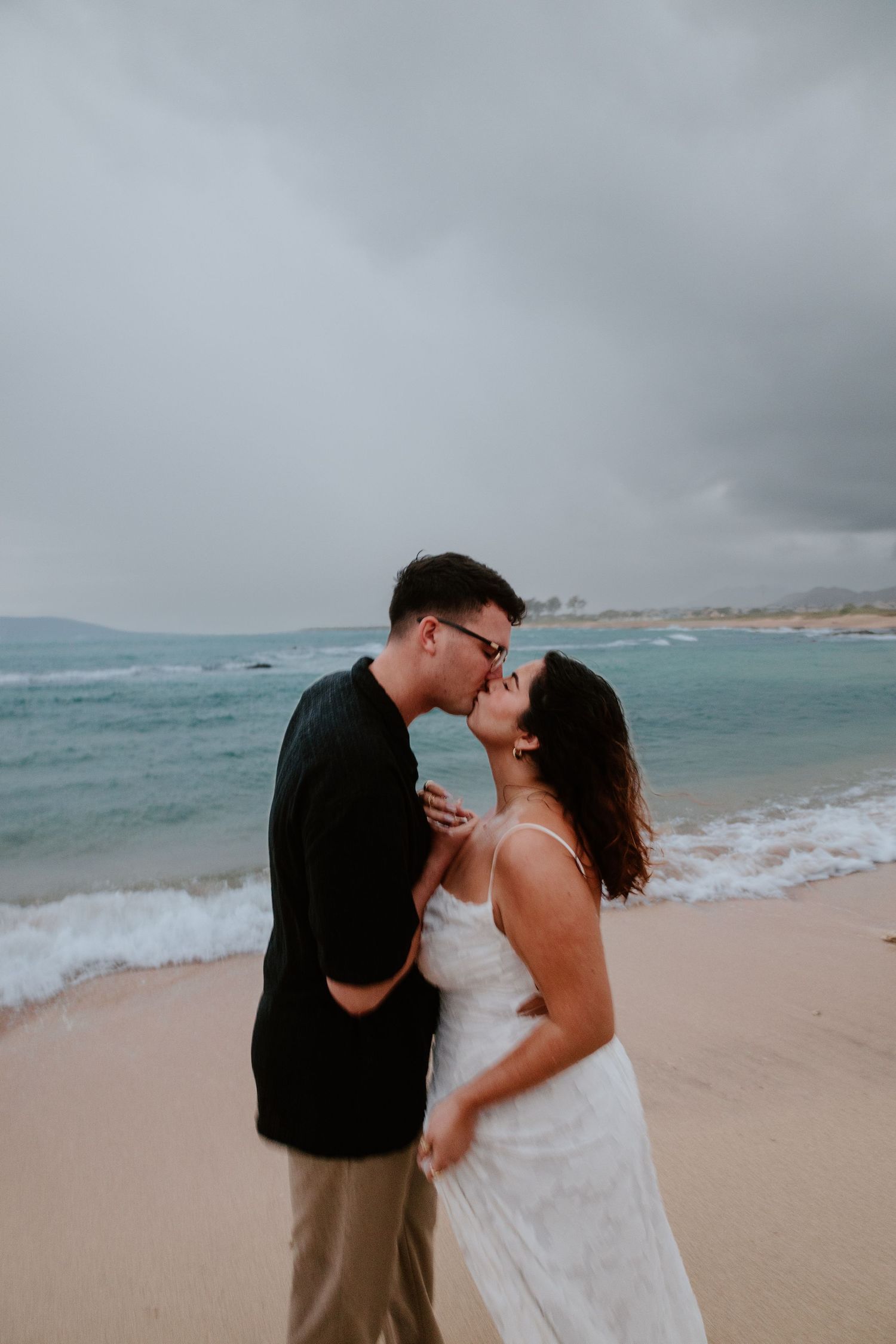 A bride and groom share a romantic kiss on a sandy beach with ocean waves under stormy skies.