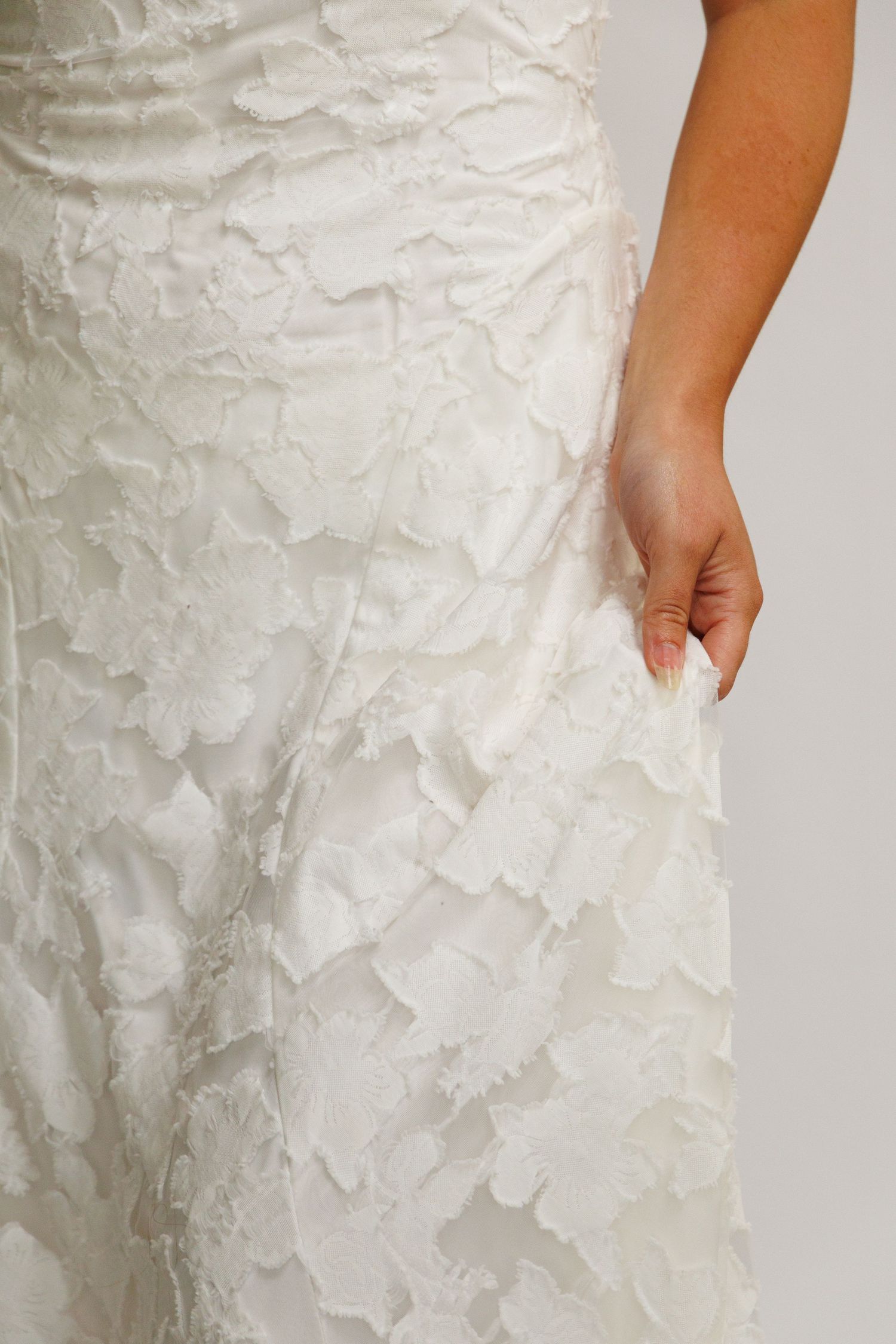 Close-up of a white floral lace wedding gown with textured appliqué details, held gently by the bride's hand.