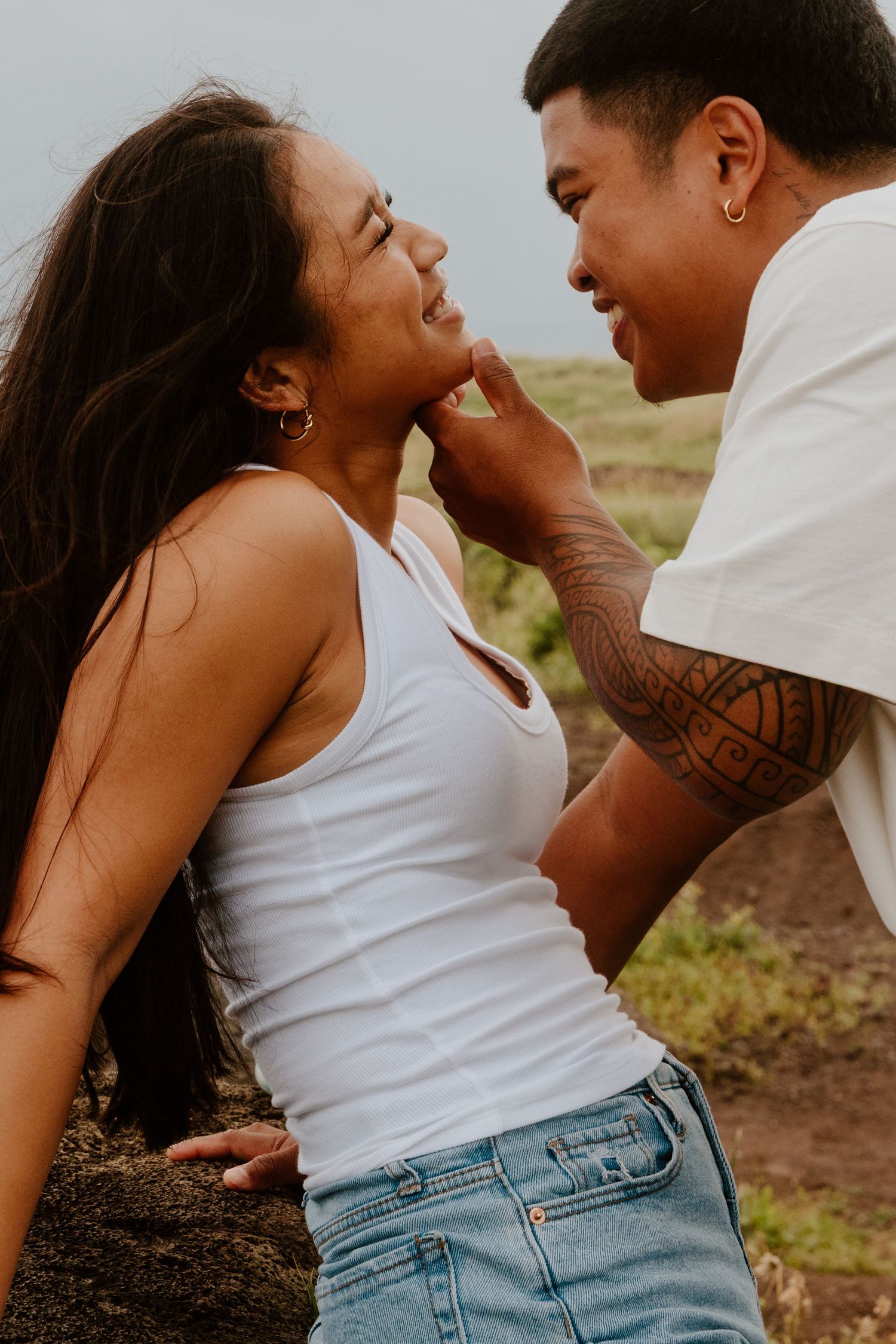 A tattooed man gently cups a smiling womans face in an intimate outdoor romantic moment.