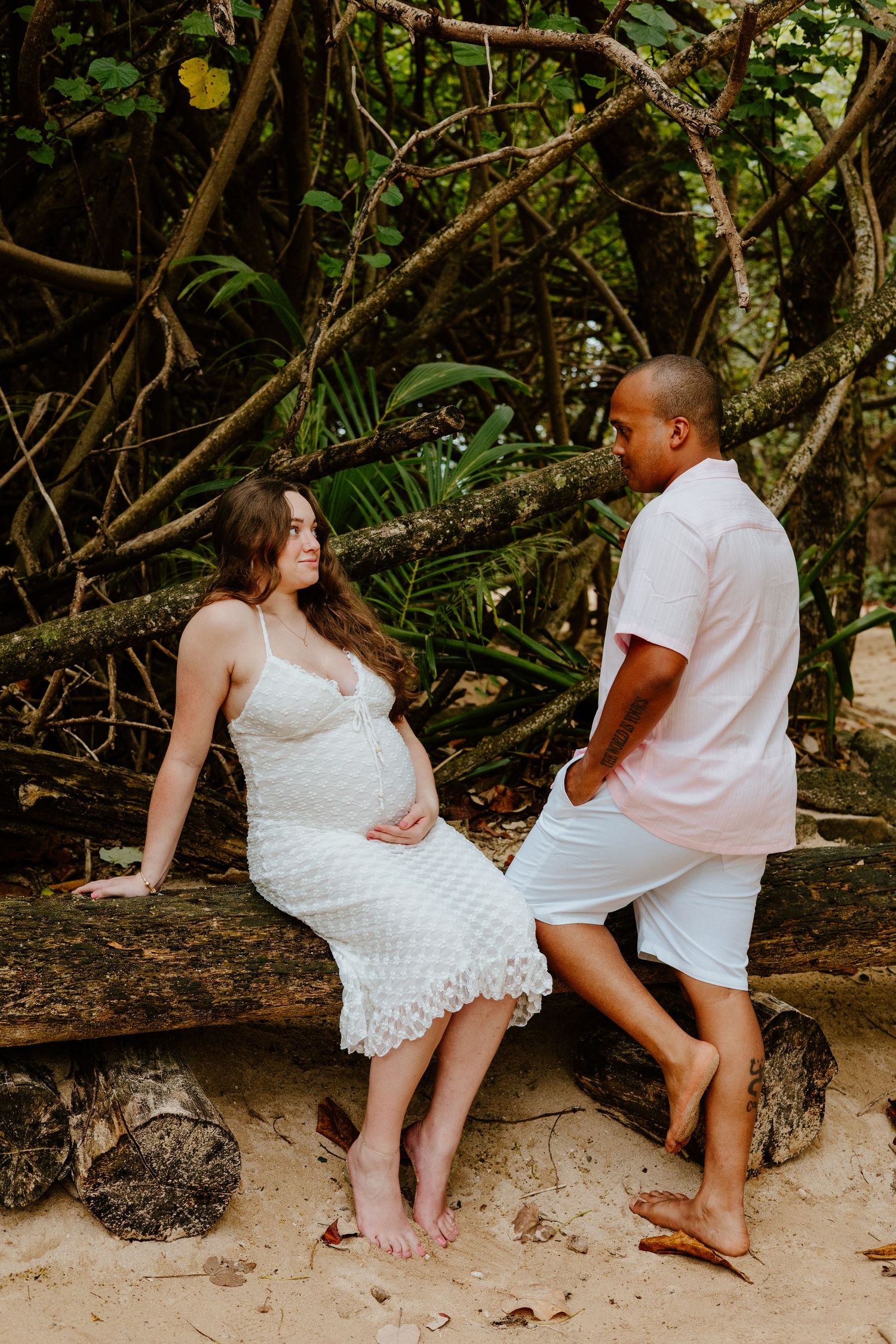 Pregnant woman in white lace dress sitting on a log with partner in a lush tropical beach setting.