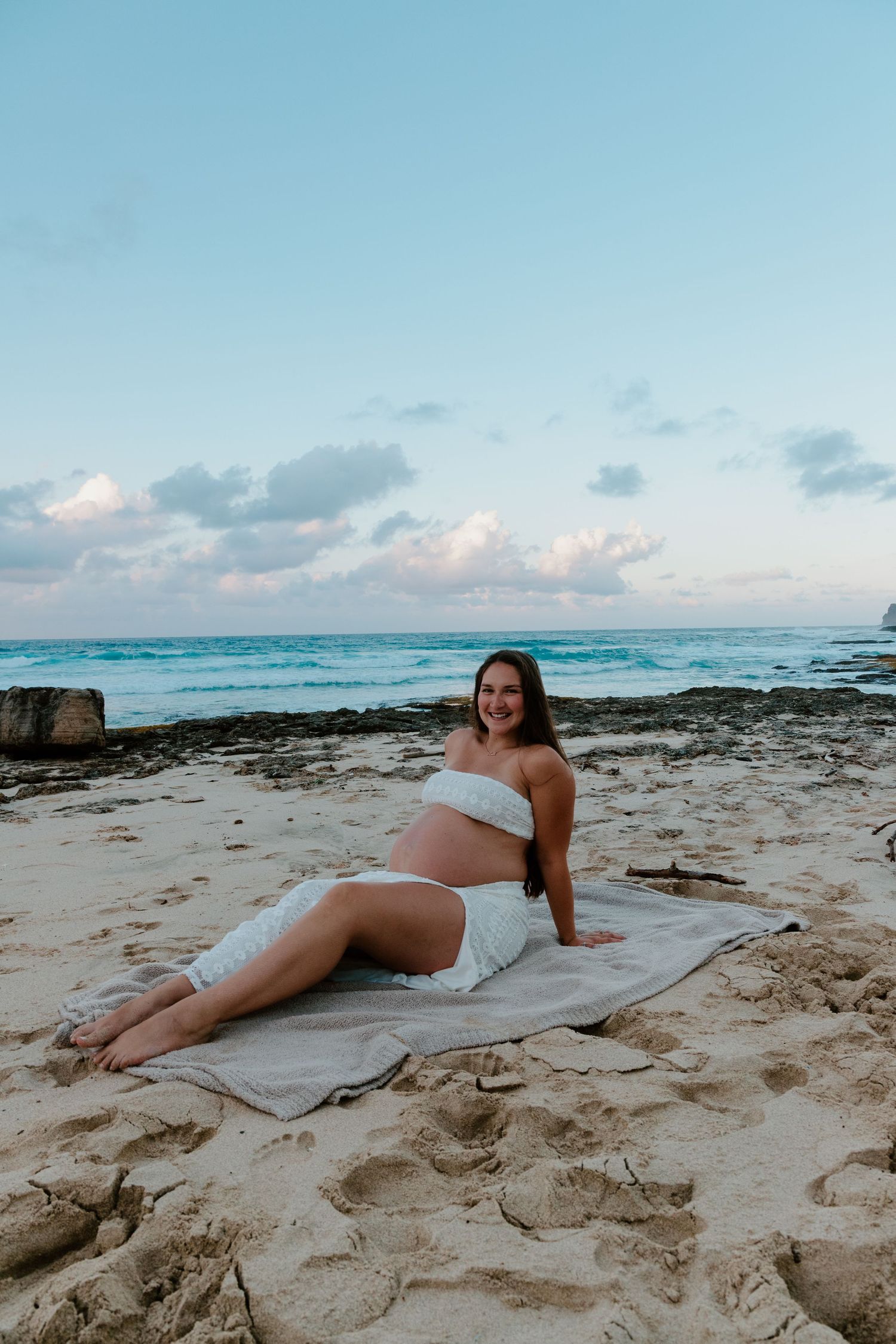 Smiling pregnant woman sitting on blanket at tropical beach during sunset.