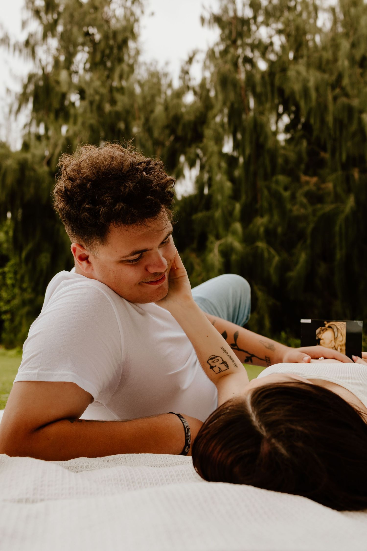 Young couple relaxing on a blanket outdoors, sharing a tender moment in lush green park.