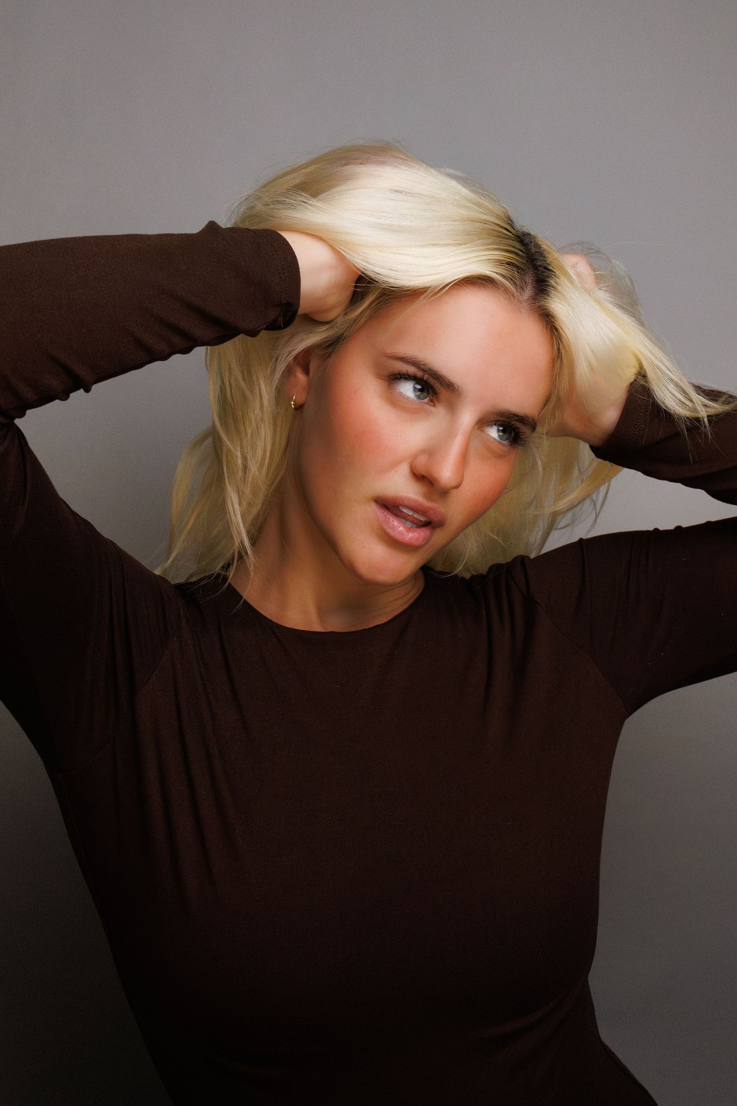 Blonde woman in brown long-sleeve top posing with hands in hair against gray background.