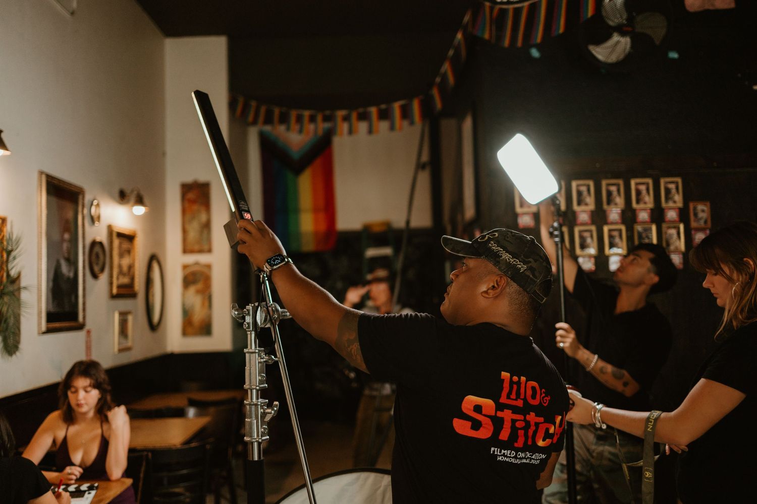 A filmmaker adjusts lighting equipment in a cozy bar with a pride flag and framed photos on the wall.