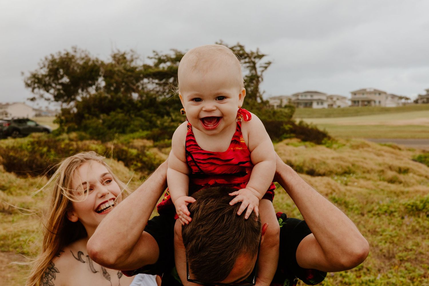Joyful family photo with parents lifting laughing baby overhead in grassy field.