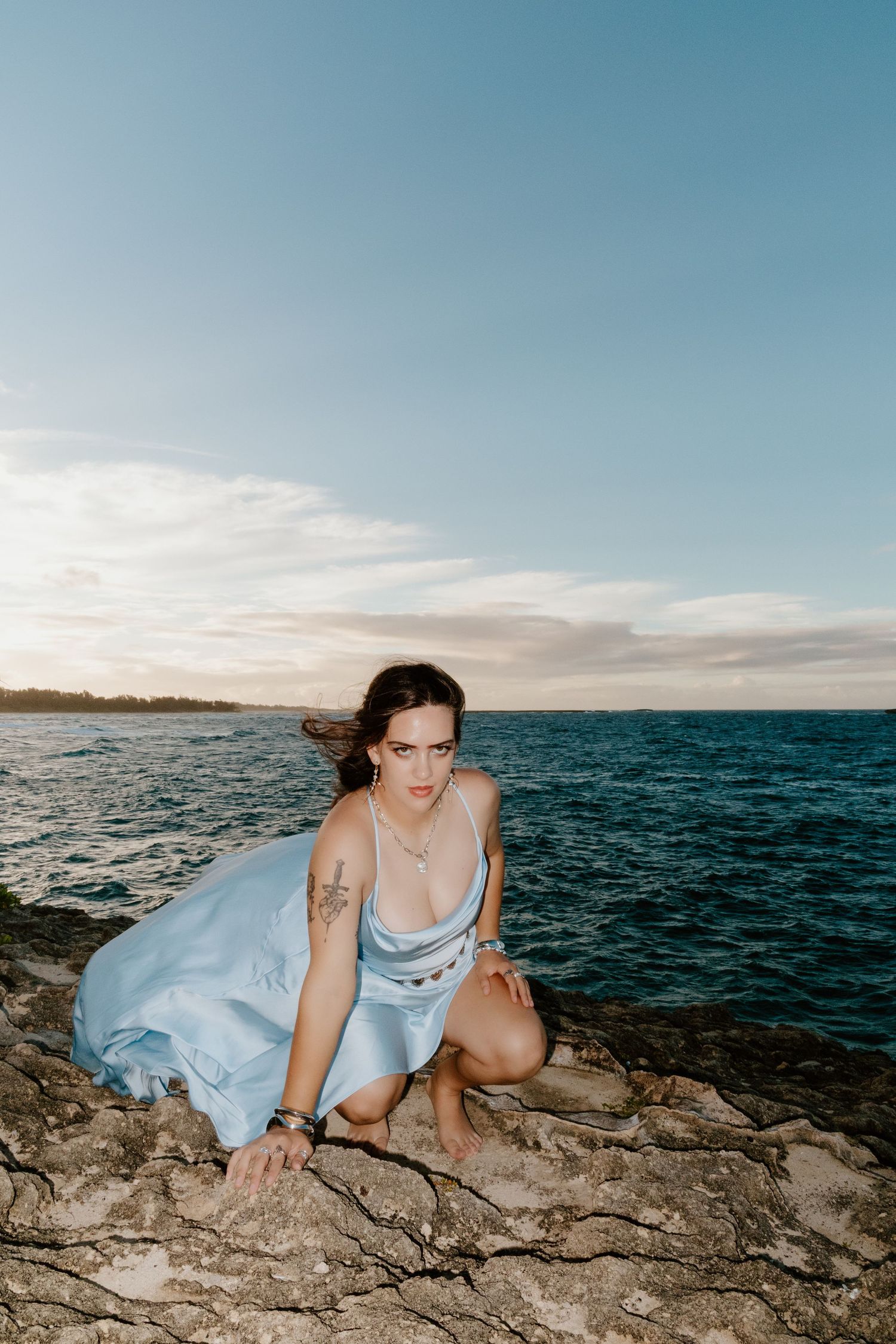Woman in light blue dress crouching on rocky shoreline at sunset with ocean and cloudy sky in background.