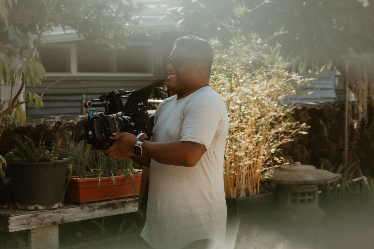 Man holding a professional camera outdoors in a sunlit garden setting near potted plants and natural foliage.