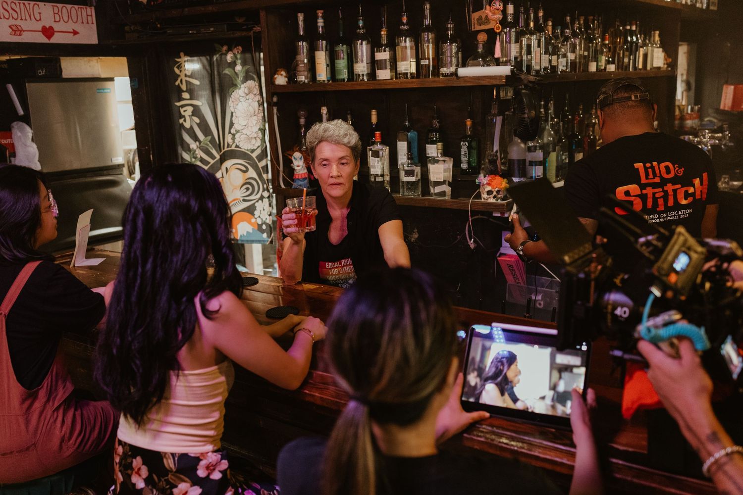 Bartender serves drinks to guests at a dark, bottle-lined bar during a lively event.
