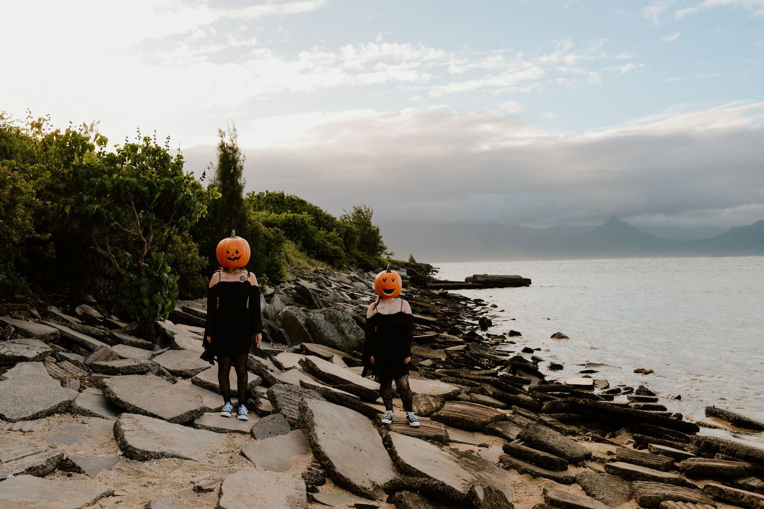 Two children wearing pumpkin masks stand on a rocky shoreline beside calm water at dusk, with green hills and a cloudy sky behind them.