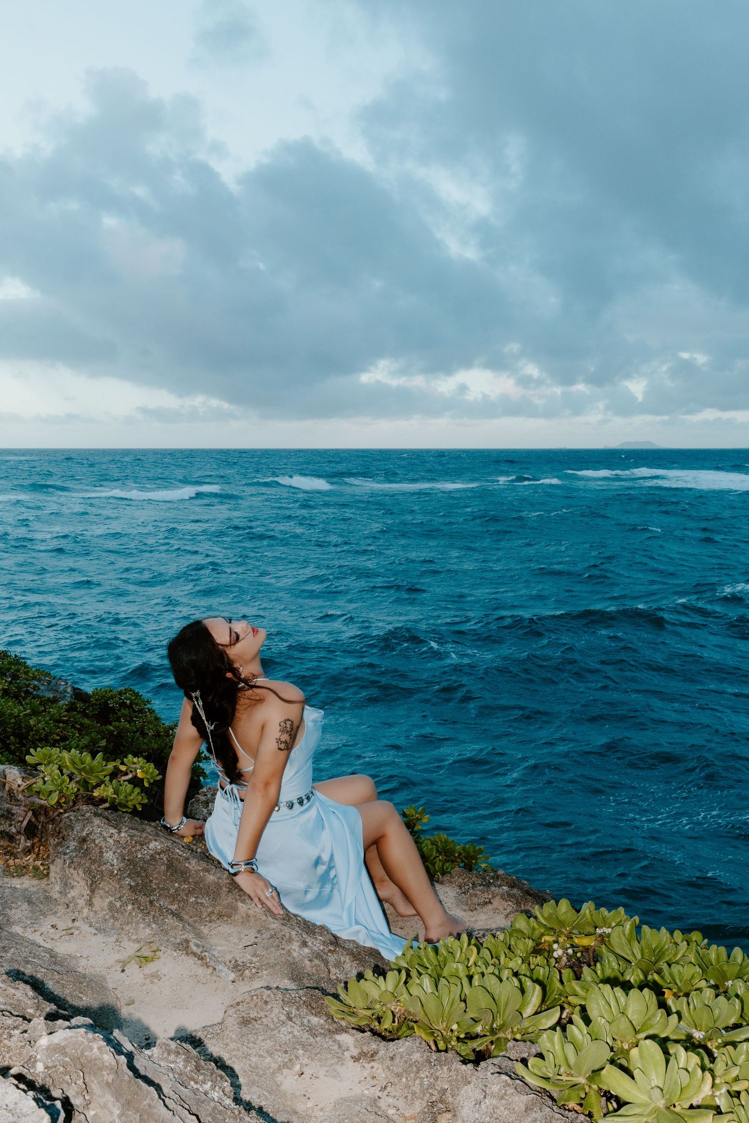 Woman in light blue dress sitting on rocky cliff overlooking turquoise ocean under cloudy sky.