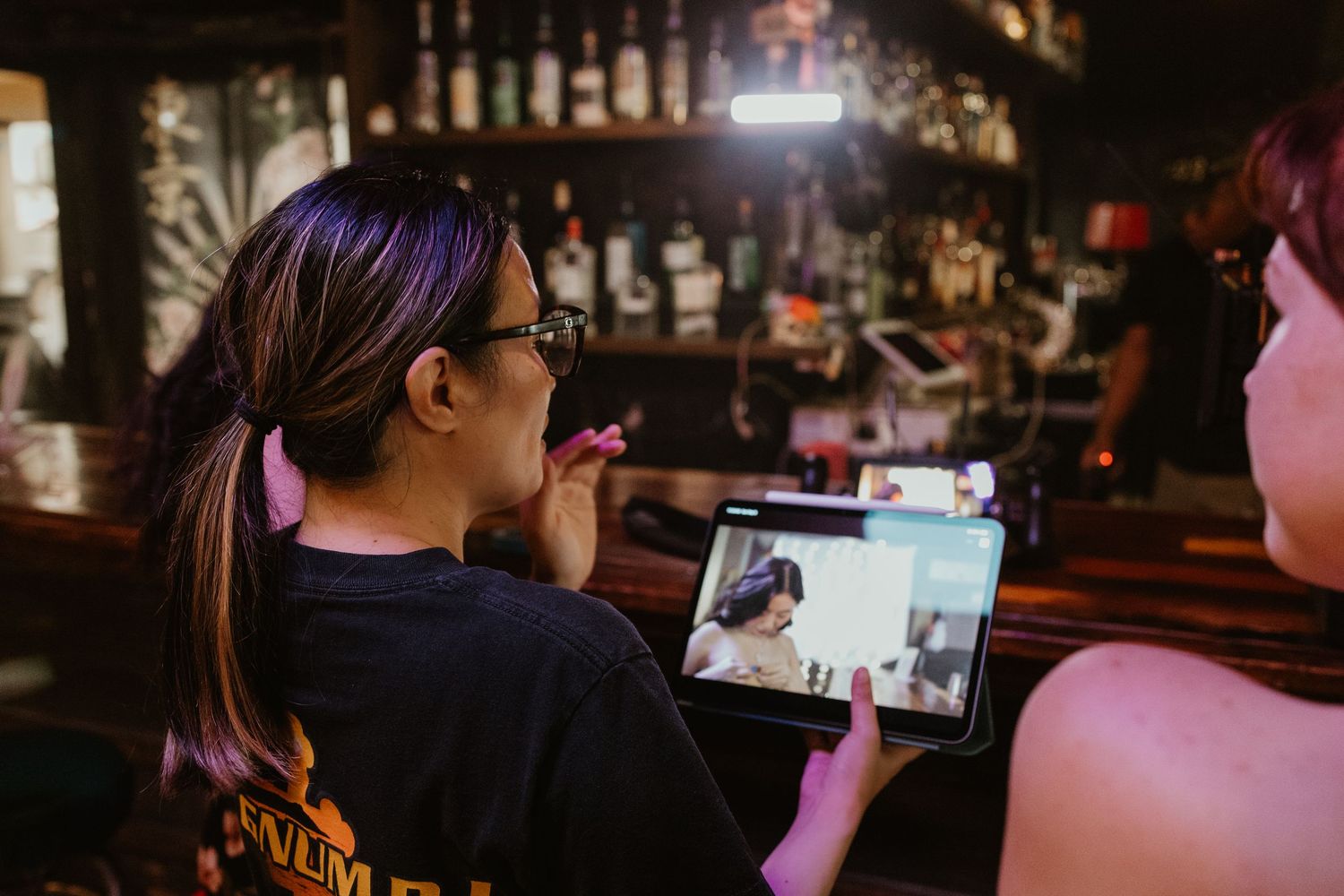 Two women at a bar look at a tablet screen together in a dimly lit venue.