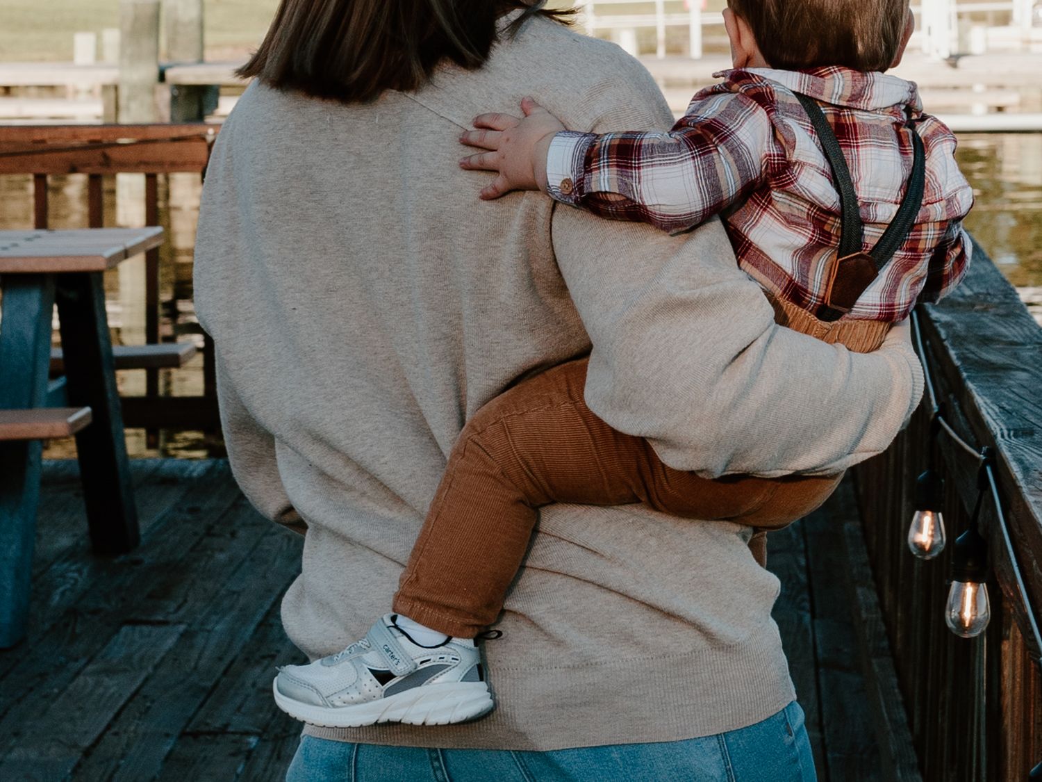 Mother in beige sweater carries toddler in plaid shirt on her hip on a wooden deck with string lights.