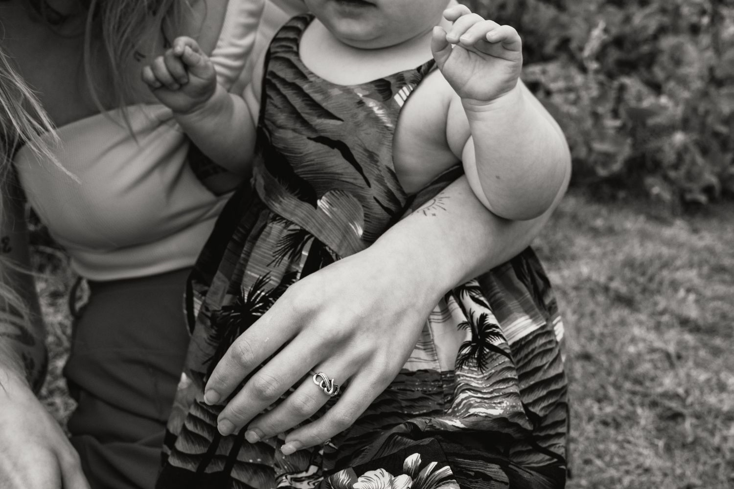 Black and white close-up of mother holding baby in floral dress with wedding ring.