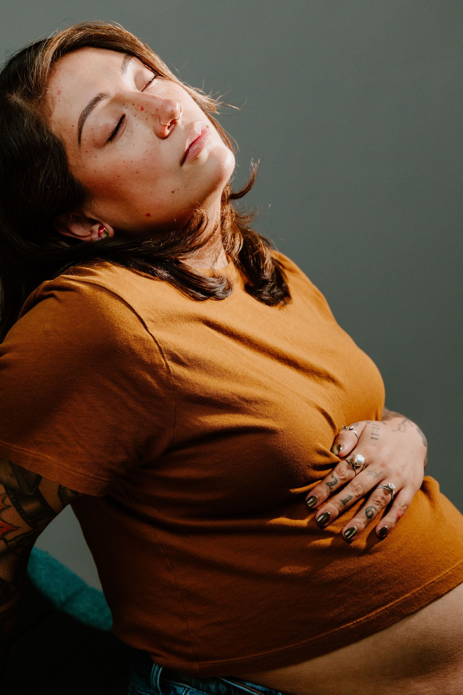 Pregnant tattooed woman in mustard yellow top holding belly with eyes closed in warm studio light.