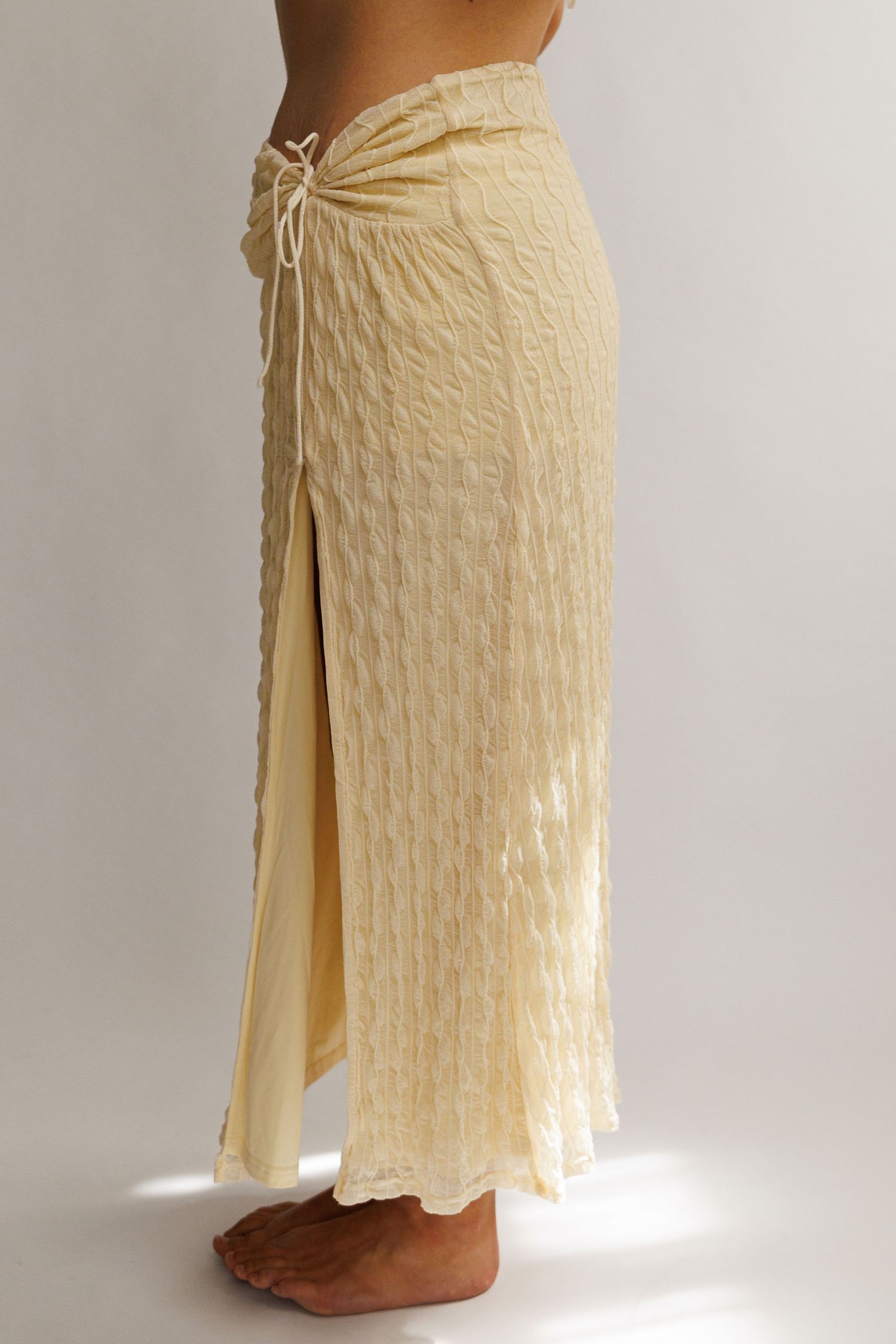 Model wearing a cream textured maxi skirt with a drawstring waist and front slit, shown from the side.
